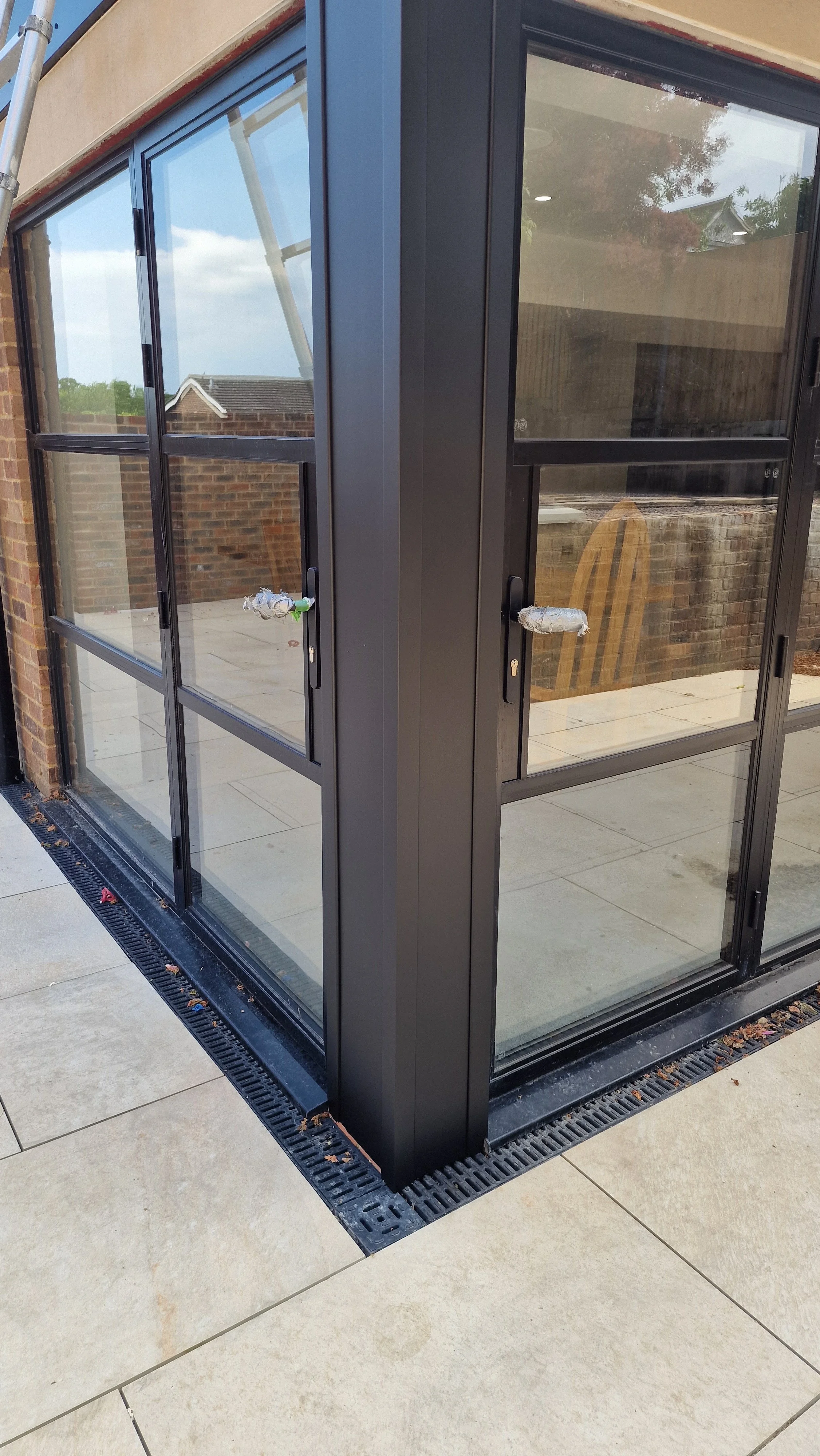 New black-framed glass sliding doors on a patio, with handles wrapped in protective plastic, and a drainage grate along the edge of the tiled floor.