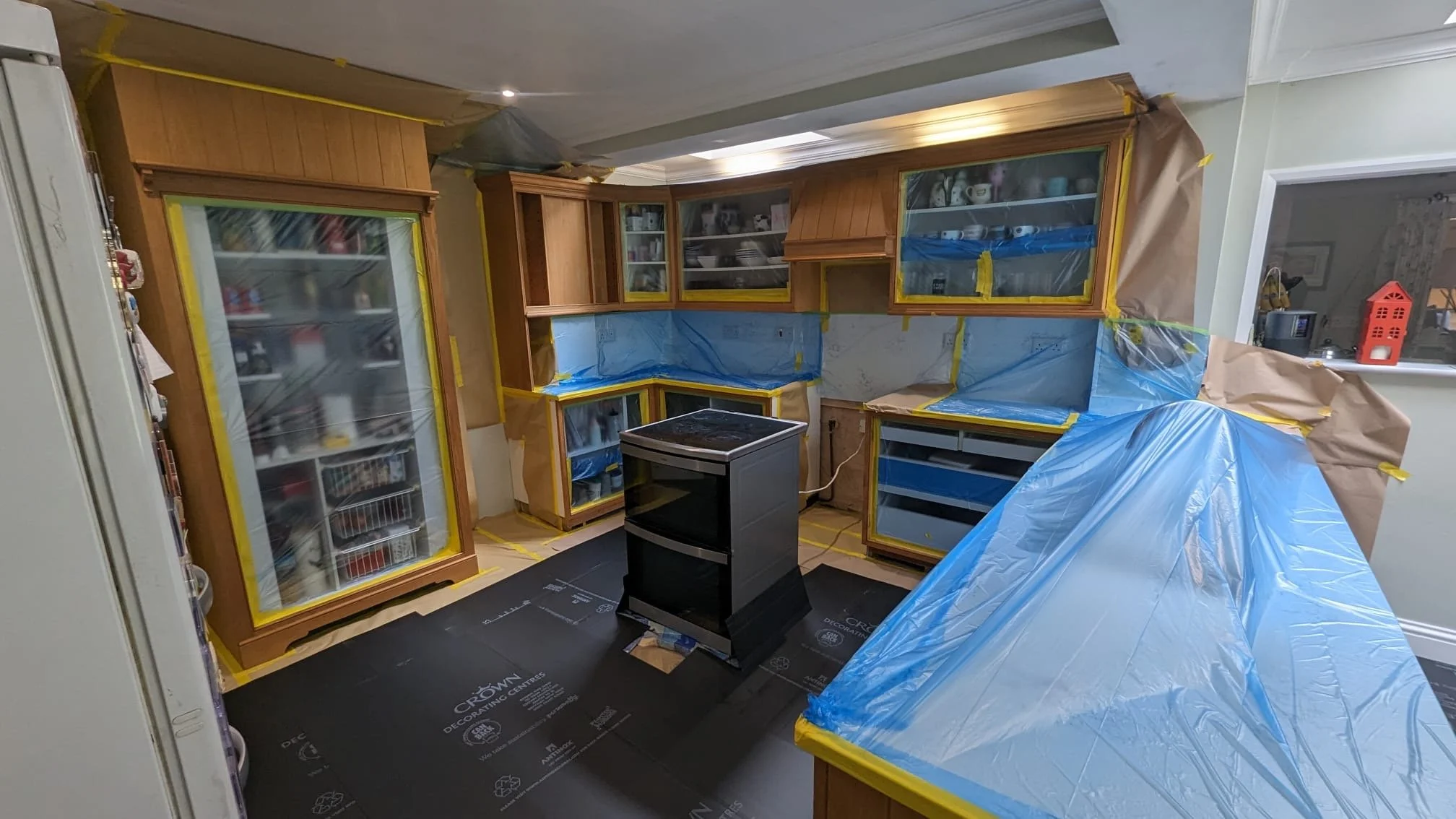 Kitchen cabinets being painted or renovated, with blue painter's tape and plastic sheeting for protection.