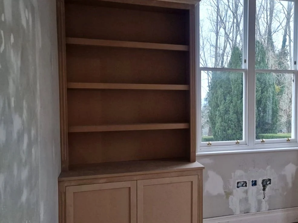 Empty wooden built-in bookshelf with four shelves next to a large window in a room under renovation.