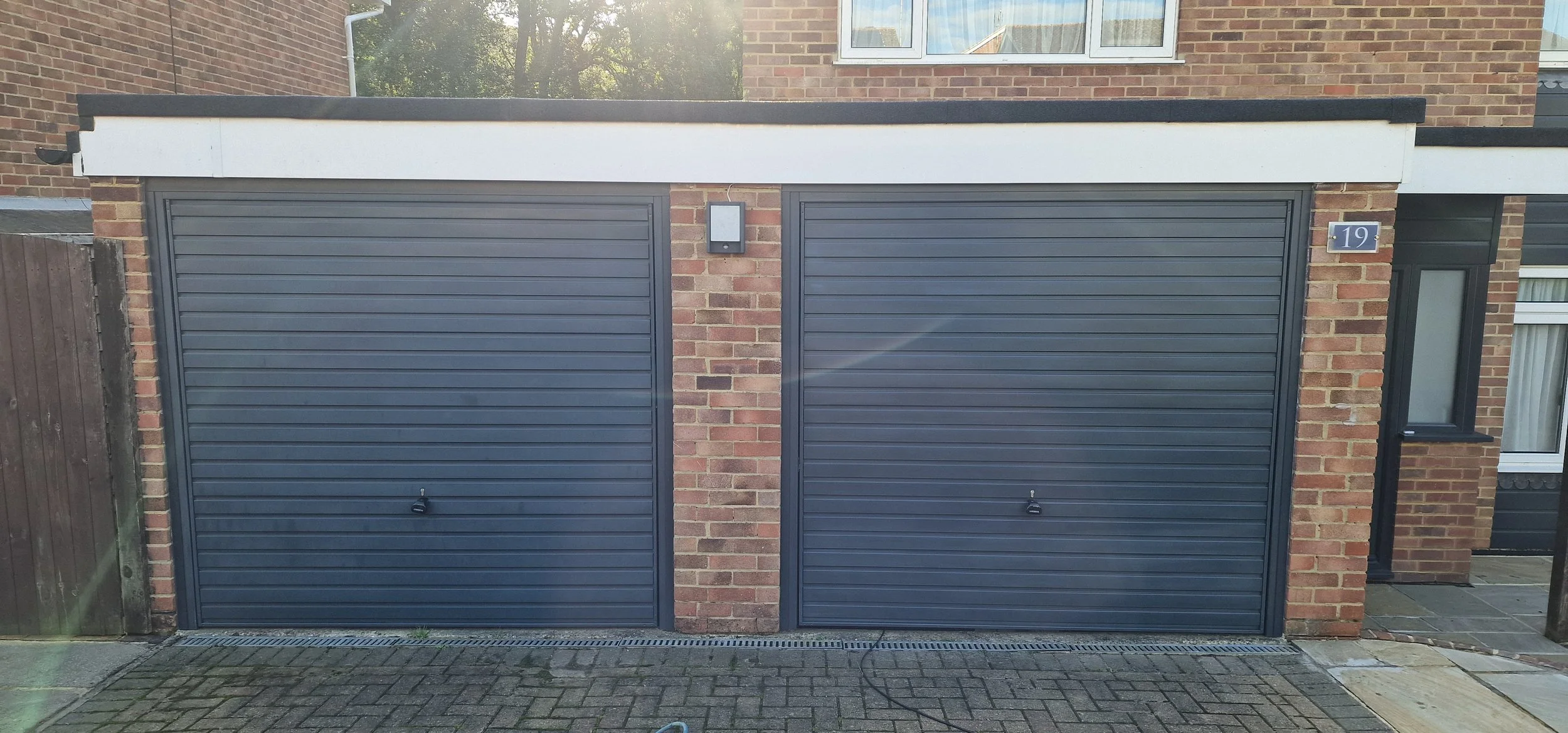 Double garage with dark gray roller doors, brick walls, a small window to the right, house number 19, and an outdoor light fixture in the center.