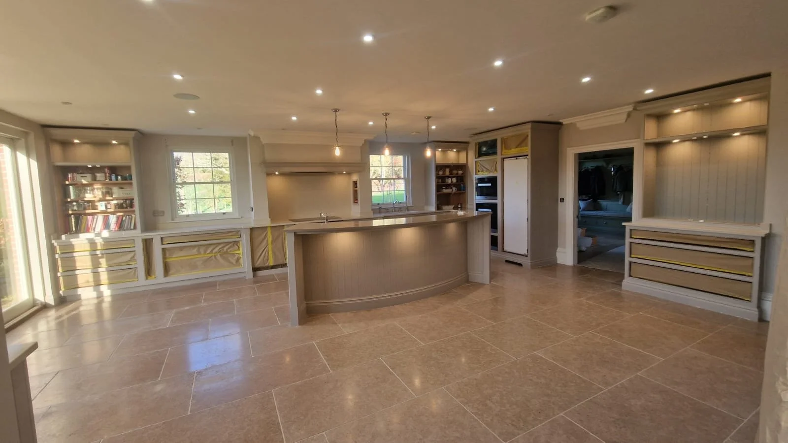 Modern kitchen with beige cabinetry, a curved island, and built-in shelves. Large windows and recessed lighting illuminate the space, with some cabinetry still wrapped and uninstalled.