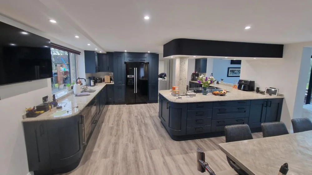 Modern kitchen with dark cabinetry, marble countertops, and a large island. Appliances include a black refrigerator, coffee maker, and toaster. There's a window with a view outside, and the room is well-lit with recessed lighting. Items like flowers and small kitchen appliances are on the counters.