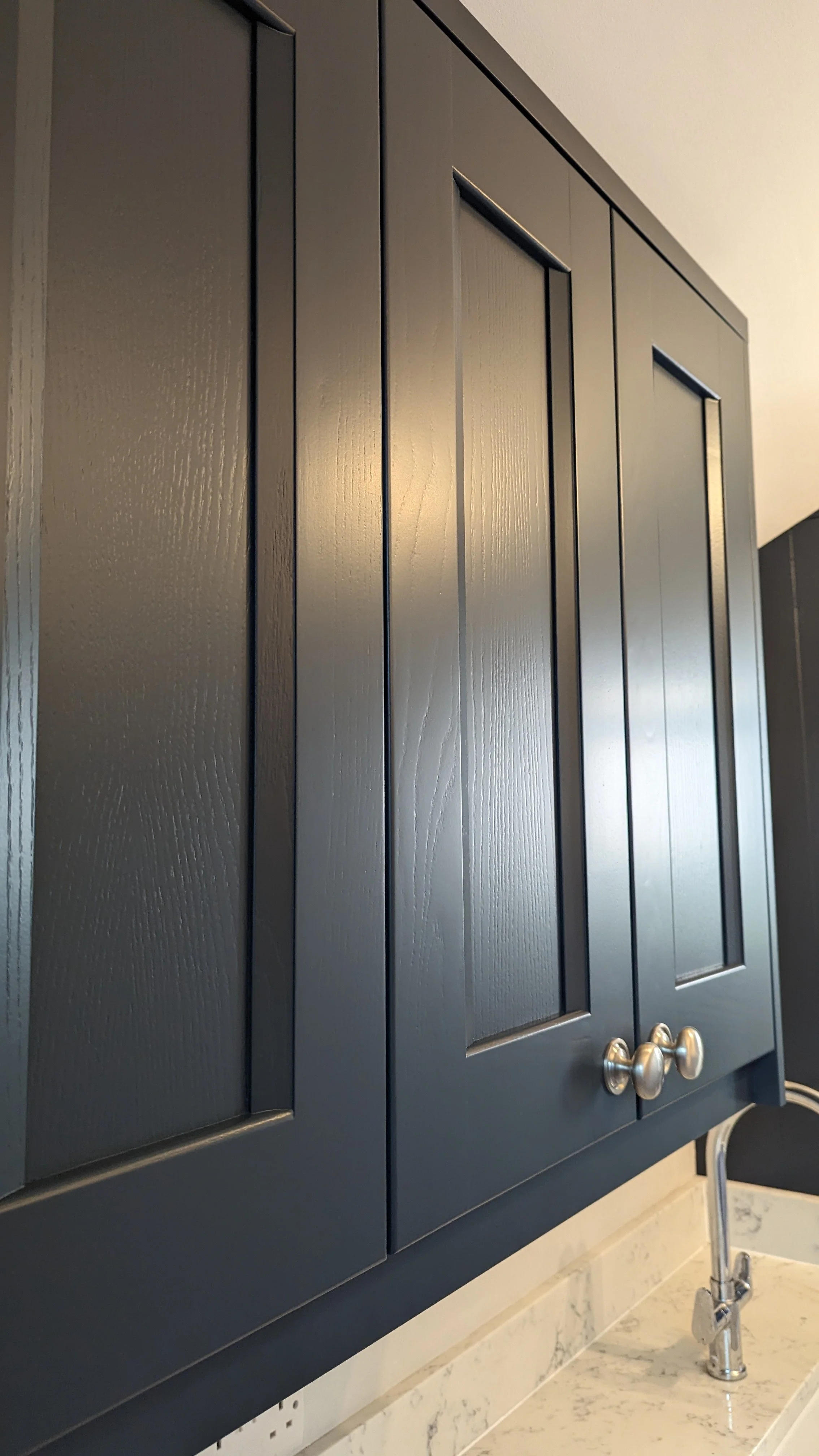 Close-up of dark grey kitchen cabinets with wood grain texture and silver knobs, and a kitchen sink with a modern chrome faucet below.