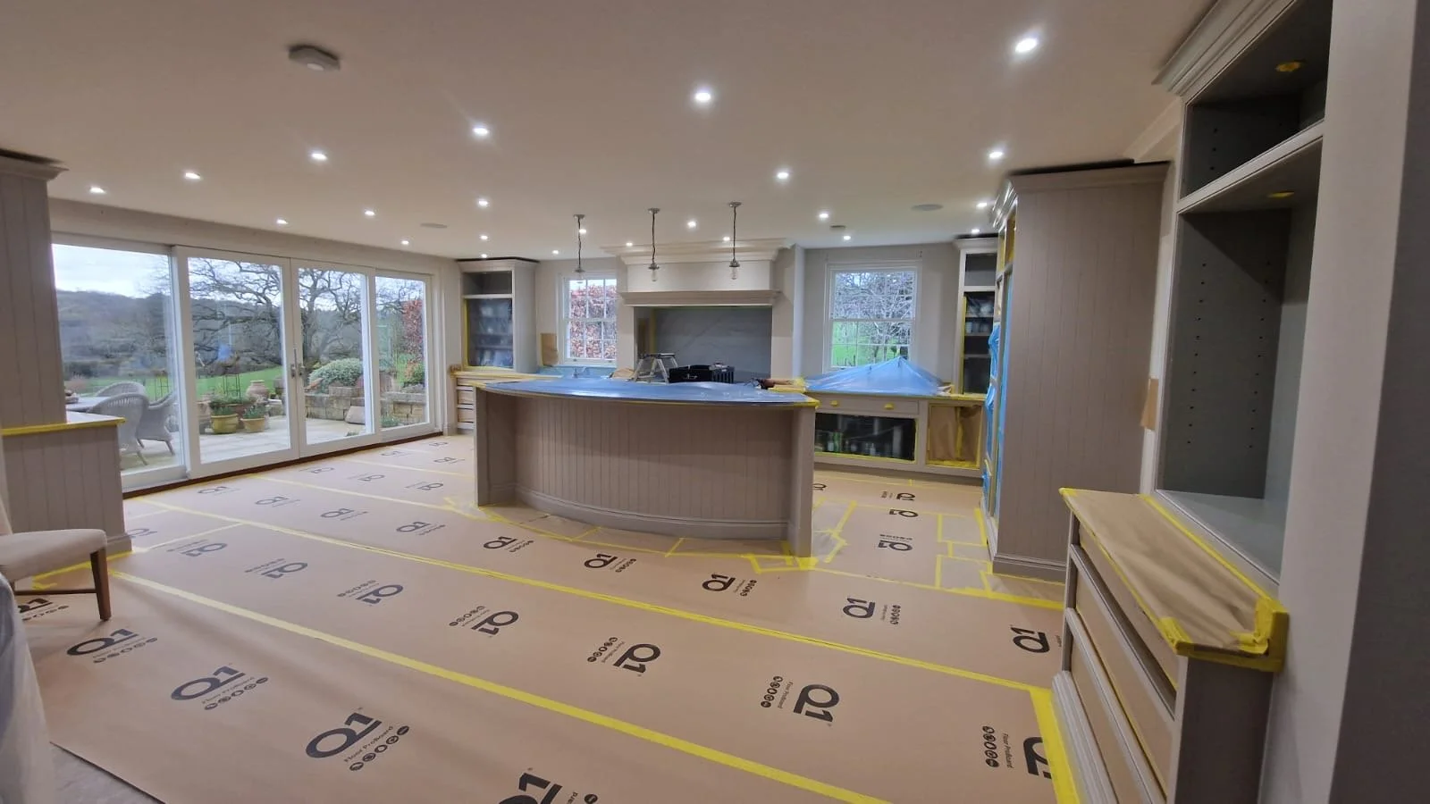 Living room under renovation with taped protective covering on the floor, cabinets, and countertops, large windows showing outdoor scenery, and ceiling lights installed.