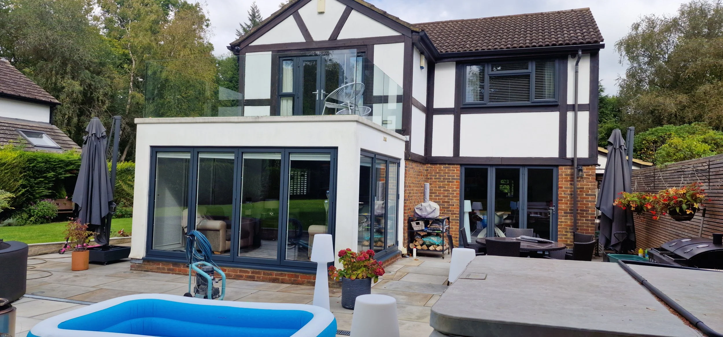 Modern two-story house with black and white exterior, glass balcony, and outdoor patio with seating, umbrellas, potted plants, and an inflatable pool.