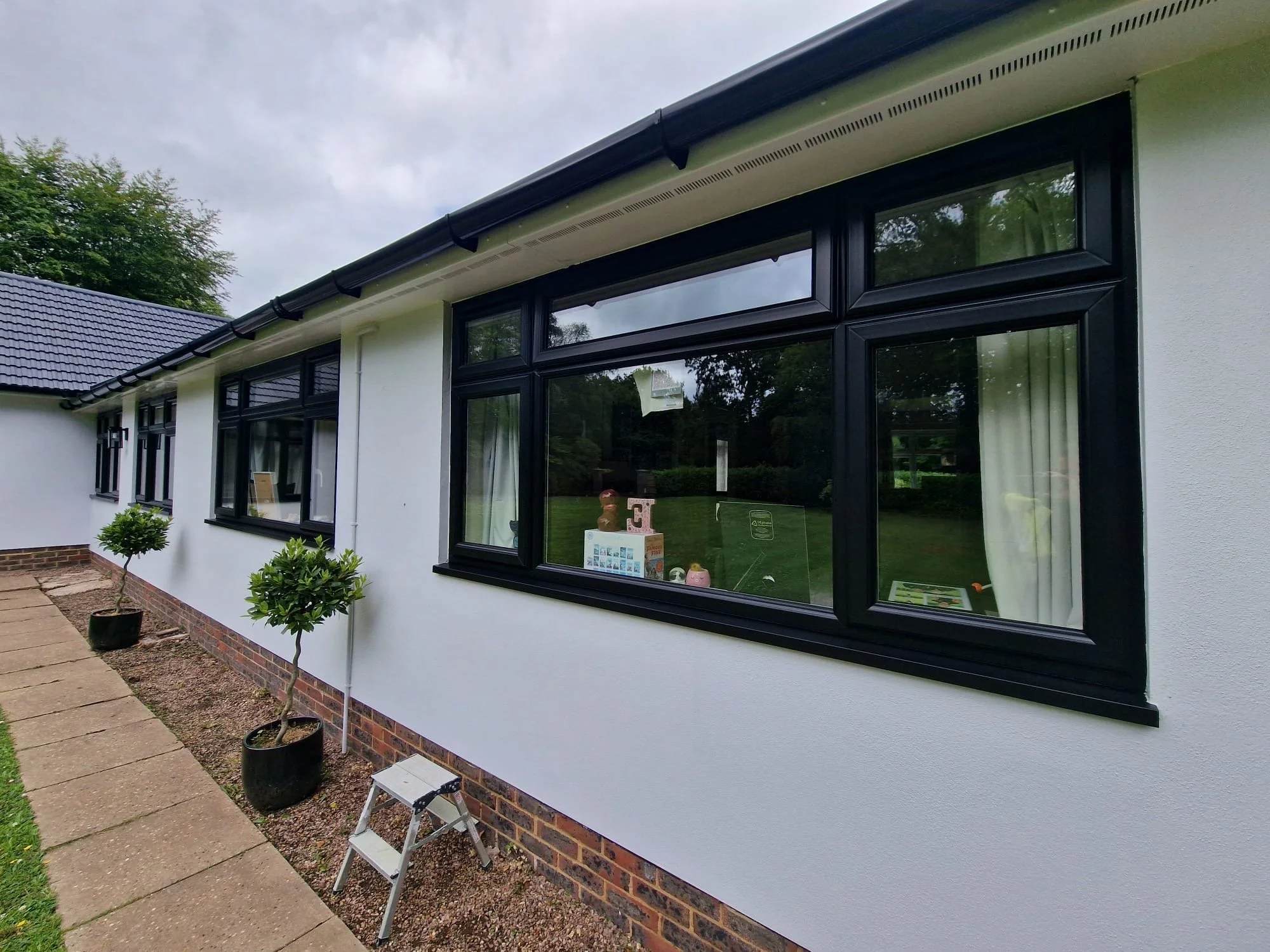 Exterior view of a white house with black-framed windows and a dark roof, two potted small trees along a stone pathway, and a cloudy sky overhead.