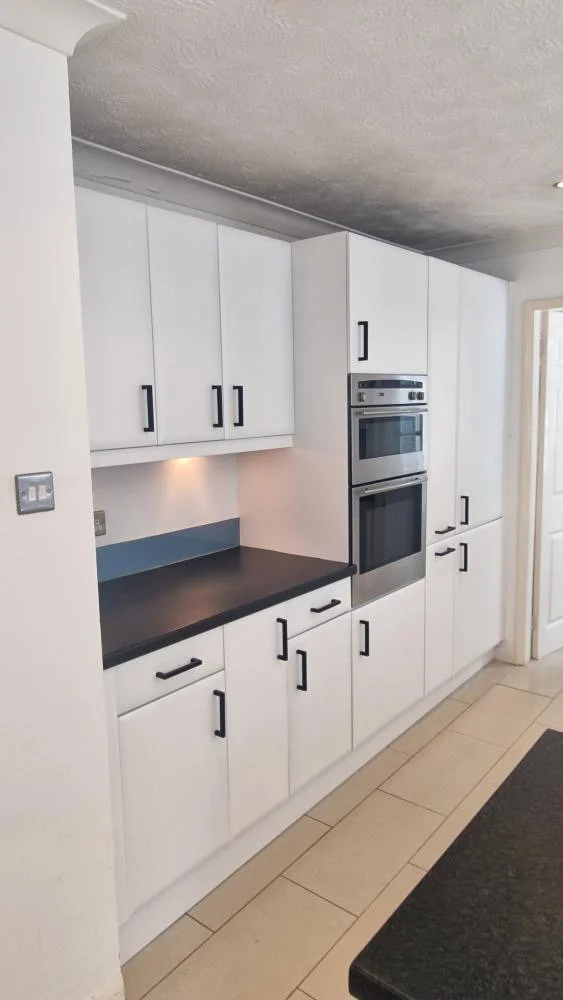 Modern kitchen with white cabinets, black handles, black countertop, built-in double oven, light-colored tiled floor, and a wall switch.