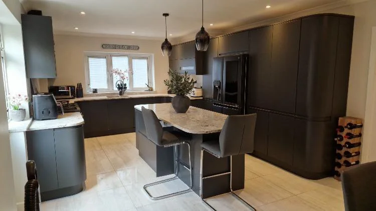 Modern kitchen with dark cabinetry, a marble island with seating, pendant lighting, and a window above the sink. Decor includes potted plants and a wine rack.