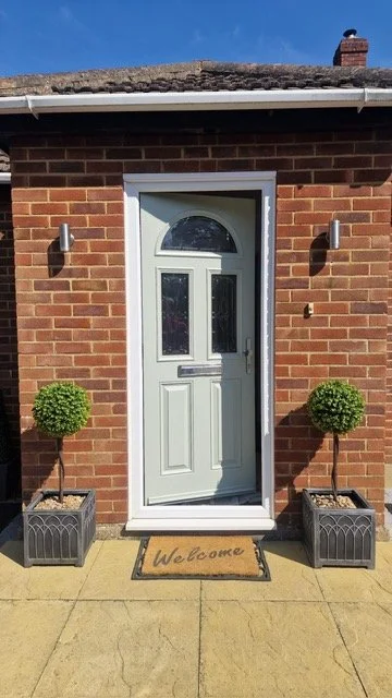 White front door with glass panels, flanked by two potted topiary plants in square planters, with wall-mounted lights on brick house exterior, and a welcome mat in front.