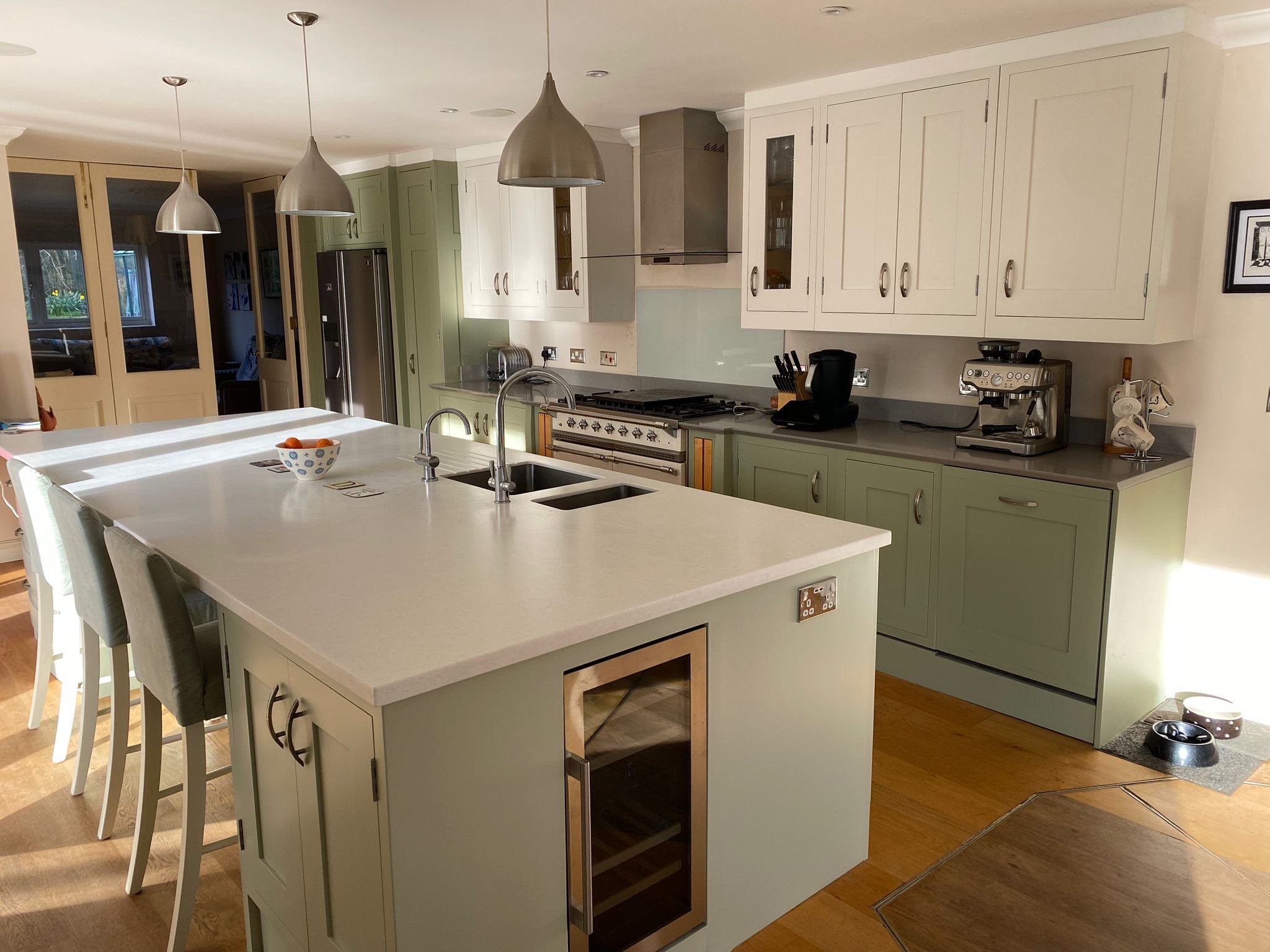 Modern kitchen with white and mint green cabinets, a large white island with a built-in wine cooler, and stainless steel appliances. There are three pendant lights hanging above the island, and two bar stools on one side. A bowl of fruit is on the island, and various kitchen appliances are on the countertop, including a coffee machine and a kettle. An area with pet bowls is visible on the floor near a window.