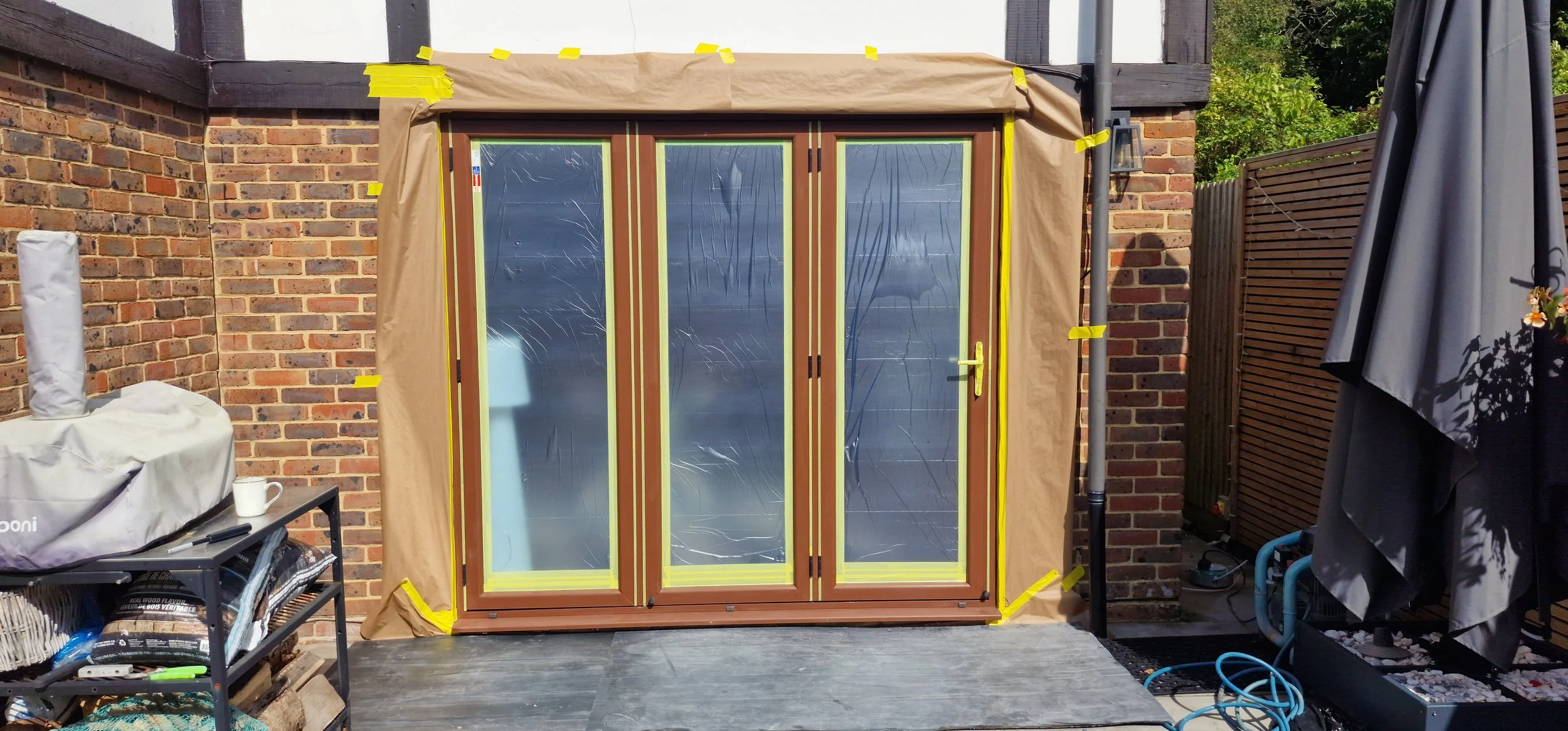 A sliding glass door with a wooden frame being installed on an outdoor patio, surrounded by masking tape and brown paper for protection.