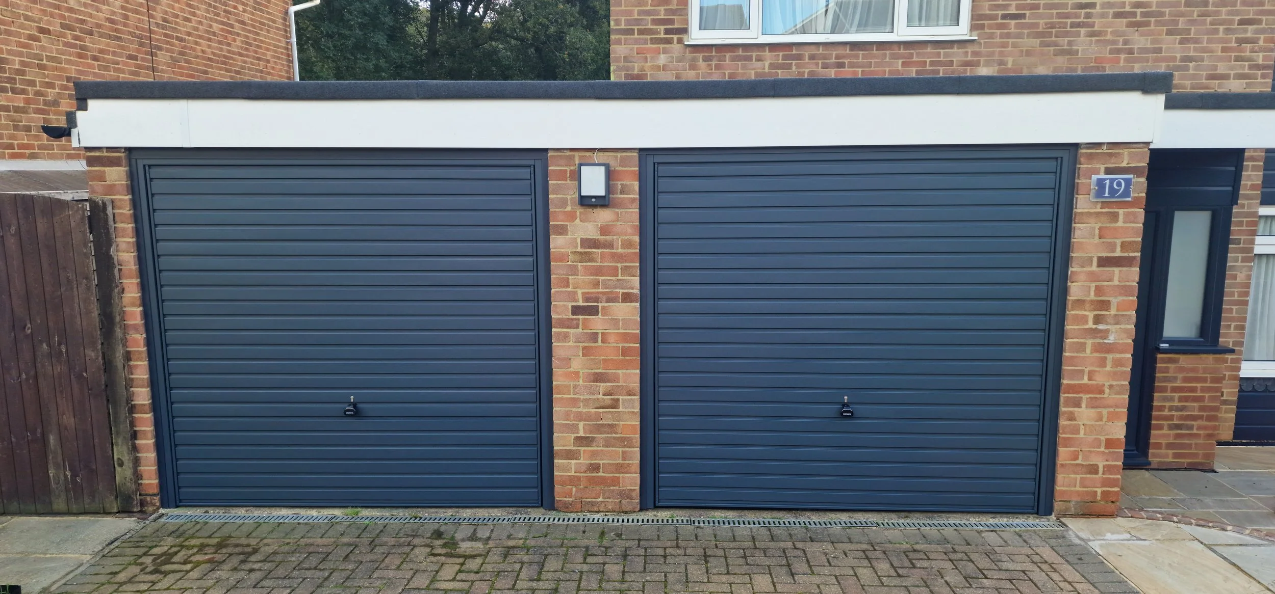 A modern brick garage with two closed blue roller shutter doors, one with a small black handle, in front of a brick driveway and a wooden fence on the left side.