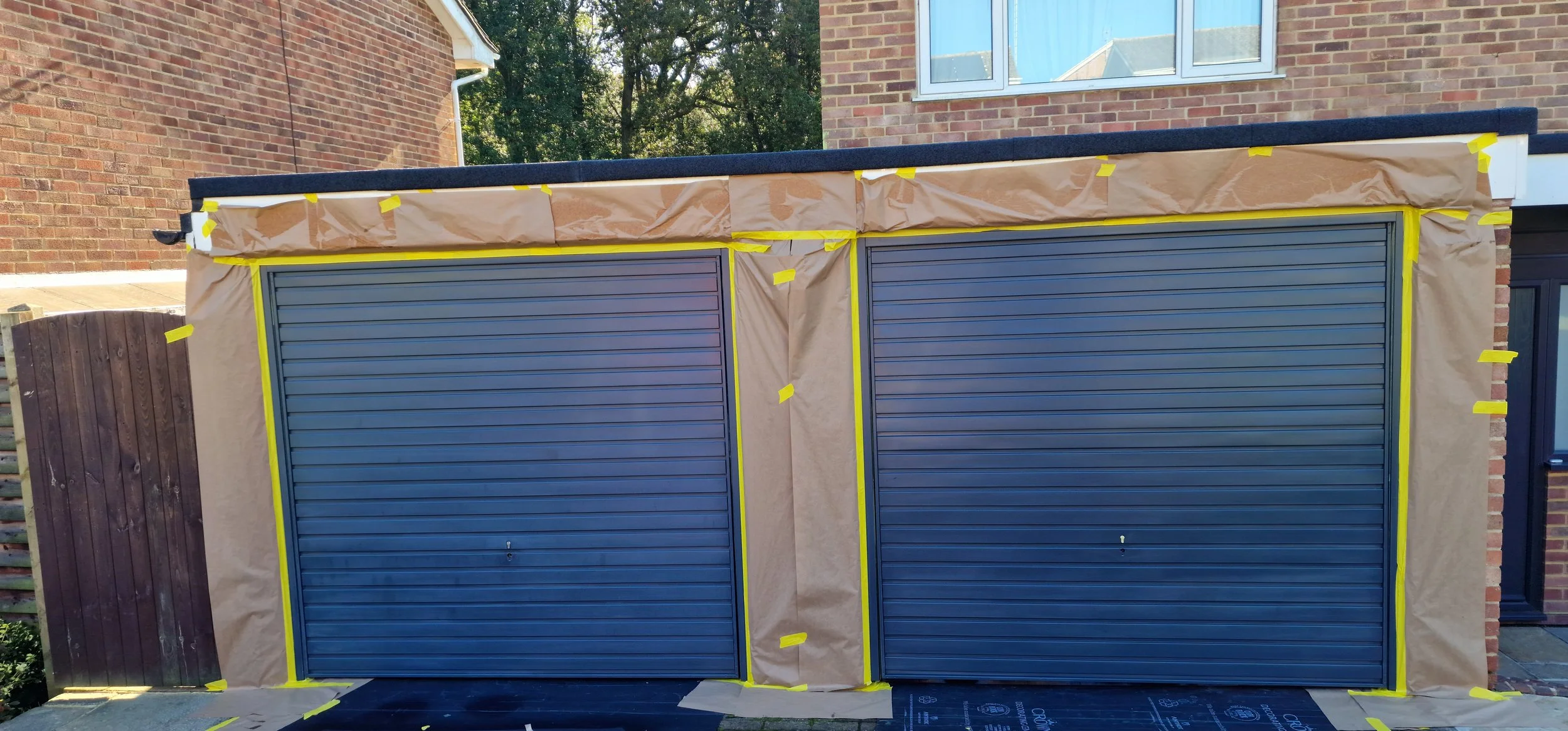 Construction site for a garage with two closed blue roller shutters, brown protective paper and yellow tape outlining the edges, brick house in the background, and a wooden fence on the left.
