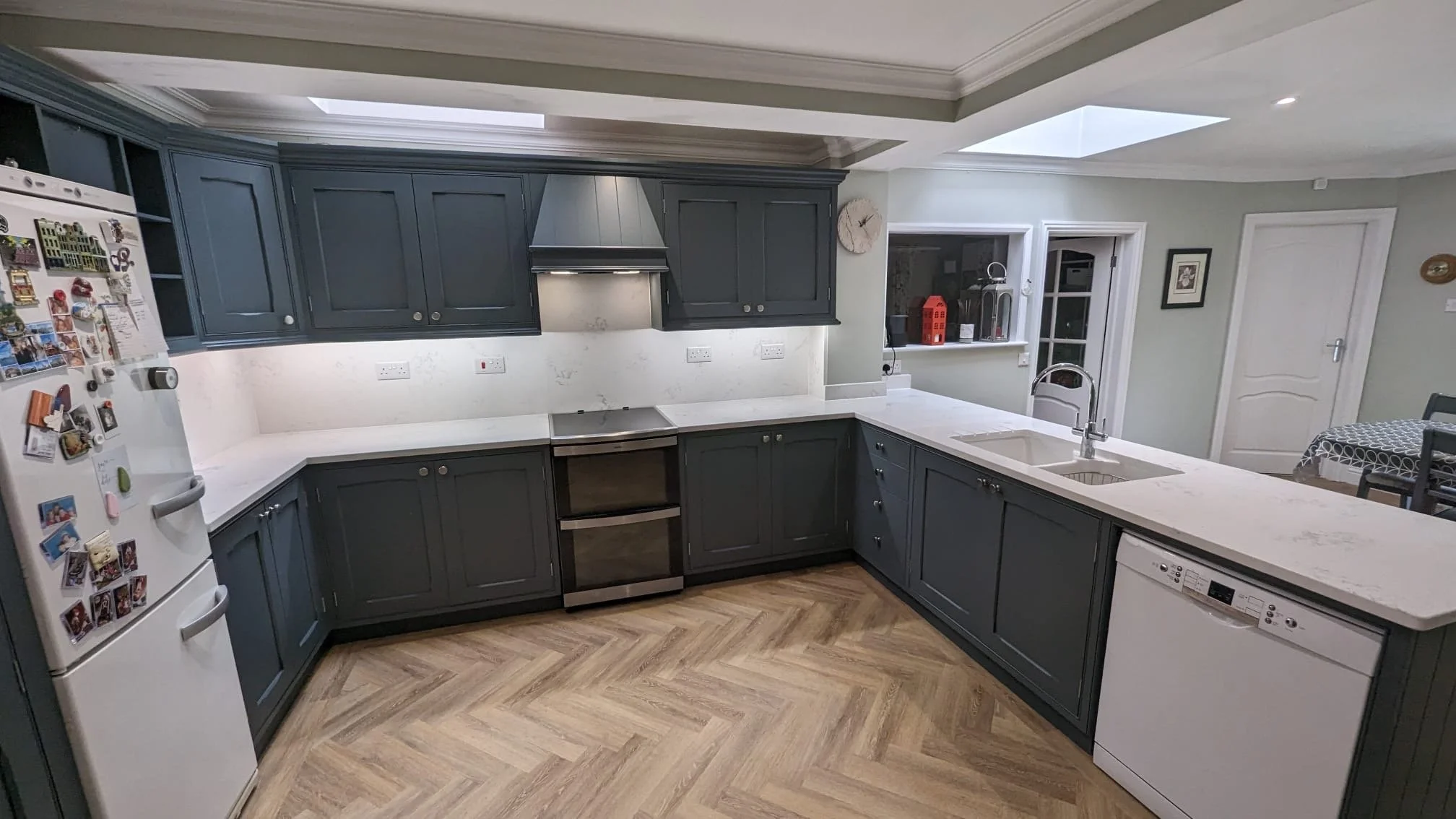 Modern kitchen with dark gray cabinets, white marble countertops, stainless steel oven and range hood, white dishwasher, white refrigerator with magnets, and a herringbone wood floor.