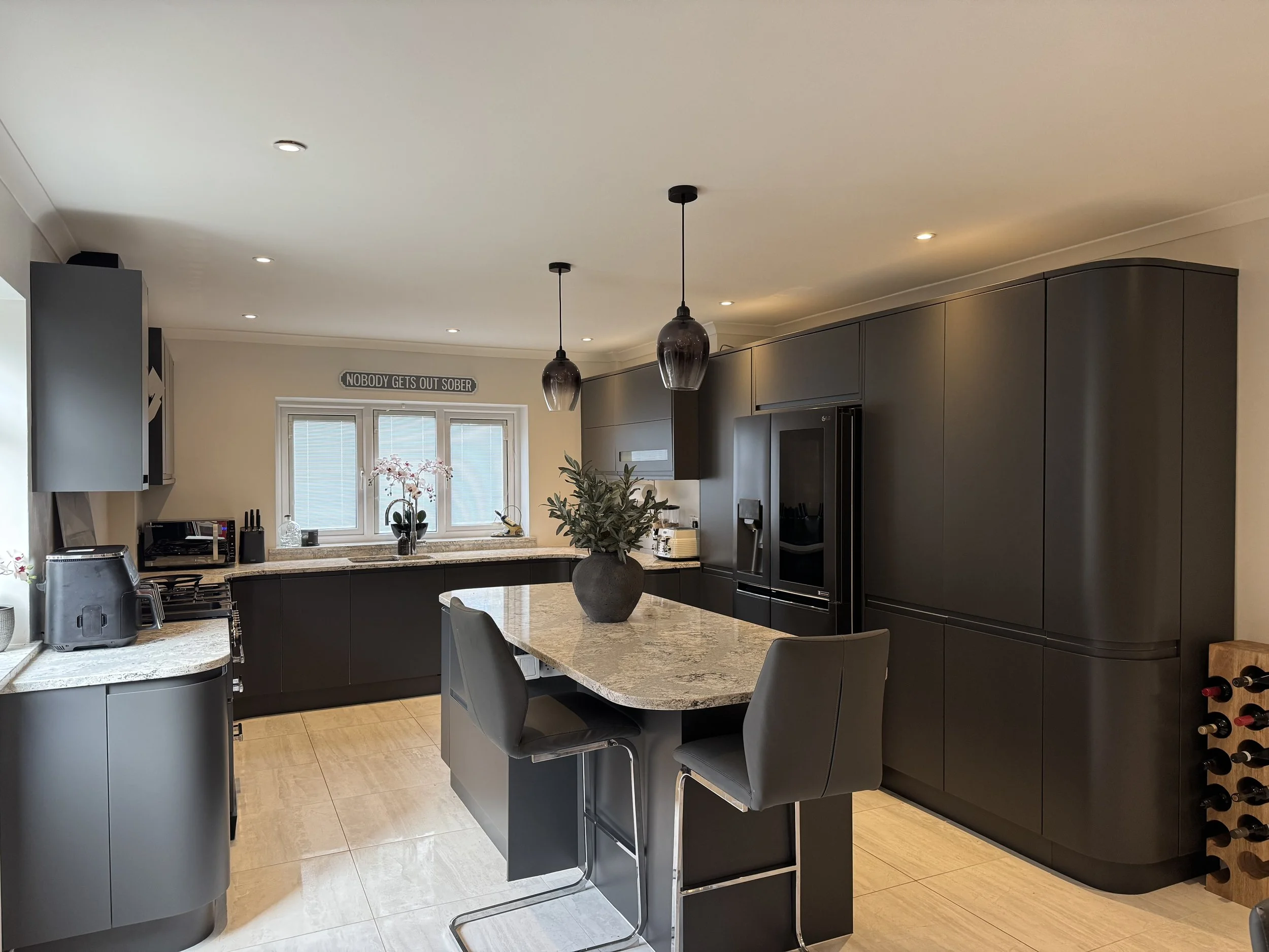 Modern kitchen with dark cabinetry, granite countertops, an island with bar stools, a window with a sign that reads 'Nobody Gets Out Sober', and various appliances and decor.