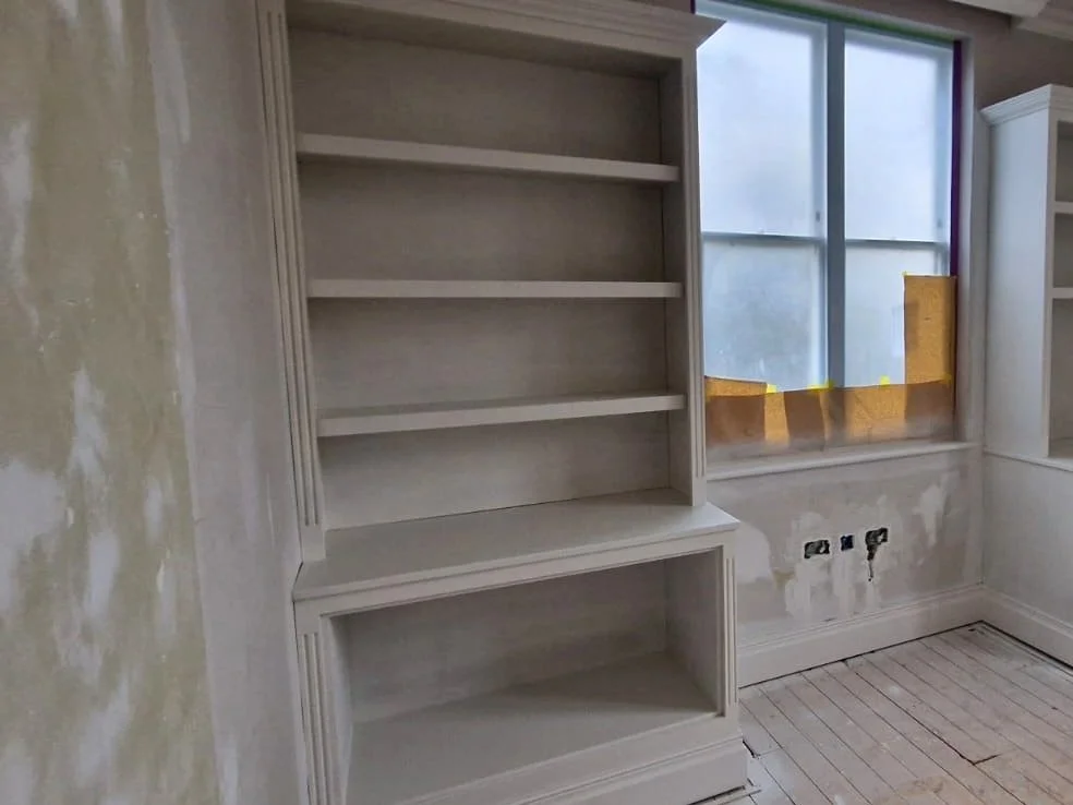 Empty built-in bookshelf with multiple shelves in a partially renovated room, with a window and unfinished wall.