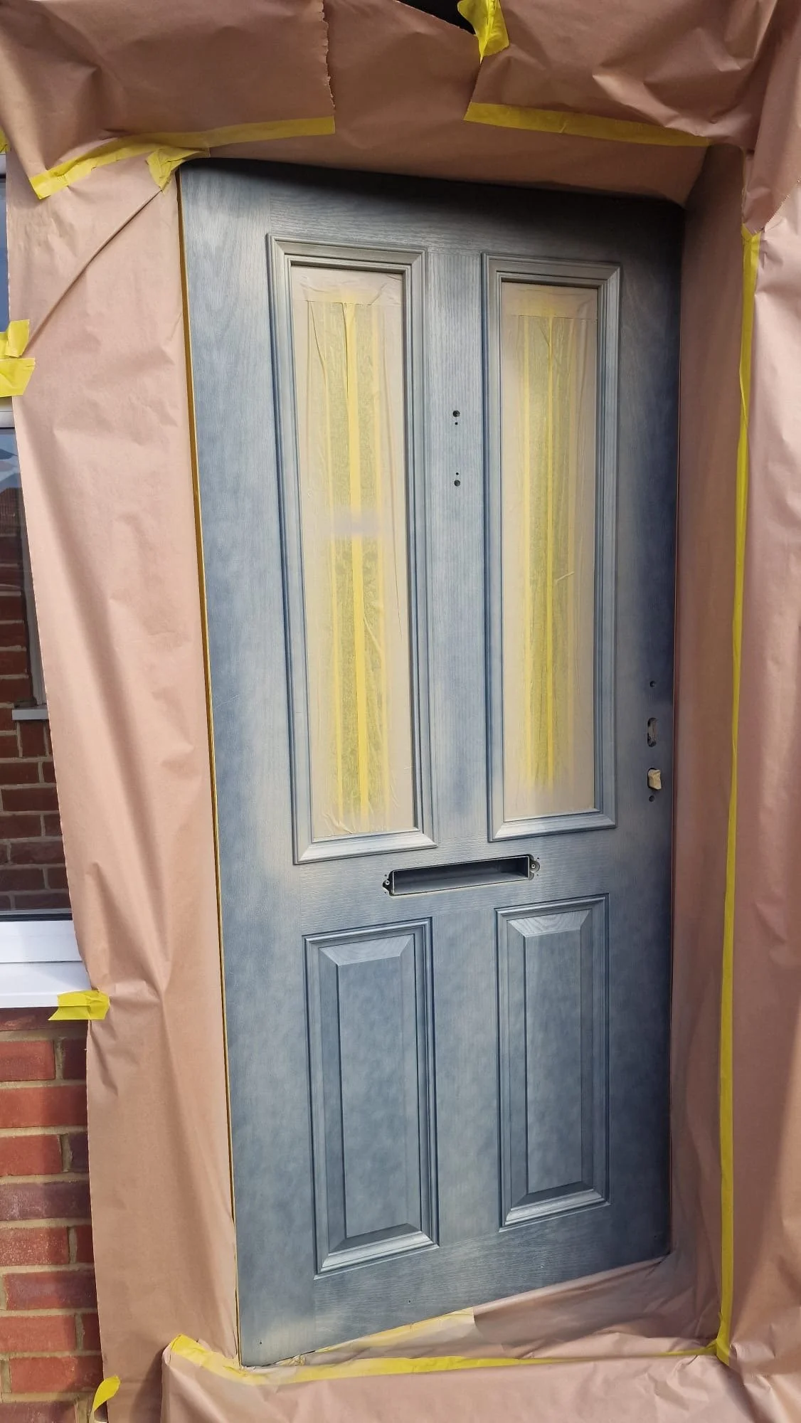 A blue front door with window cutouts, surrounded by brown masking paper with yellow tape, indicating the door is being painted or refurbished.