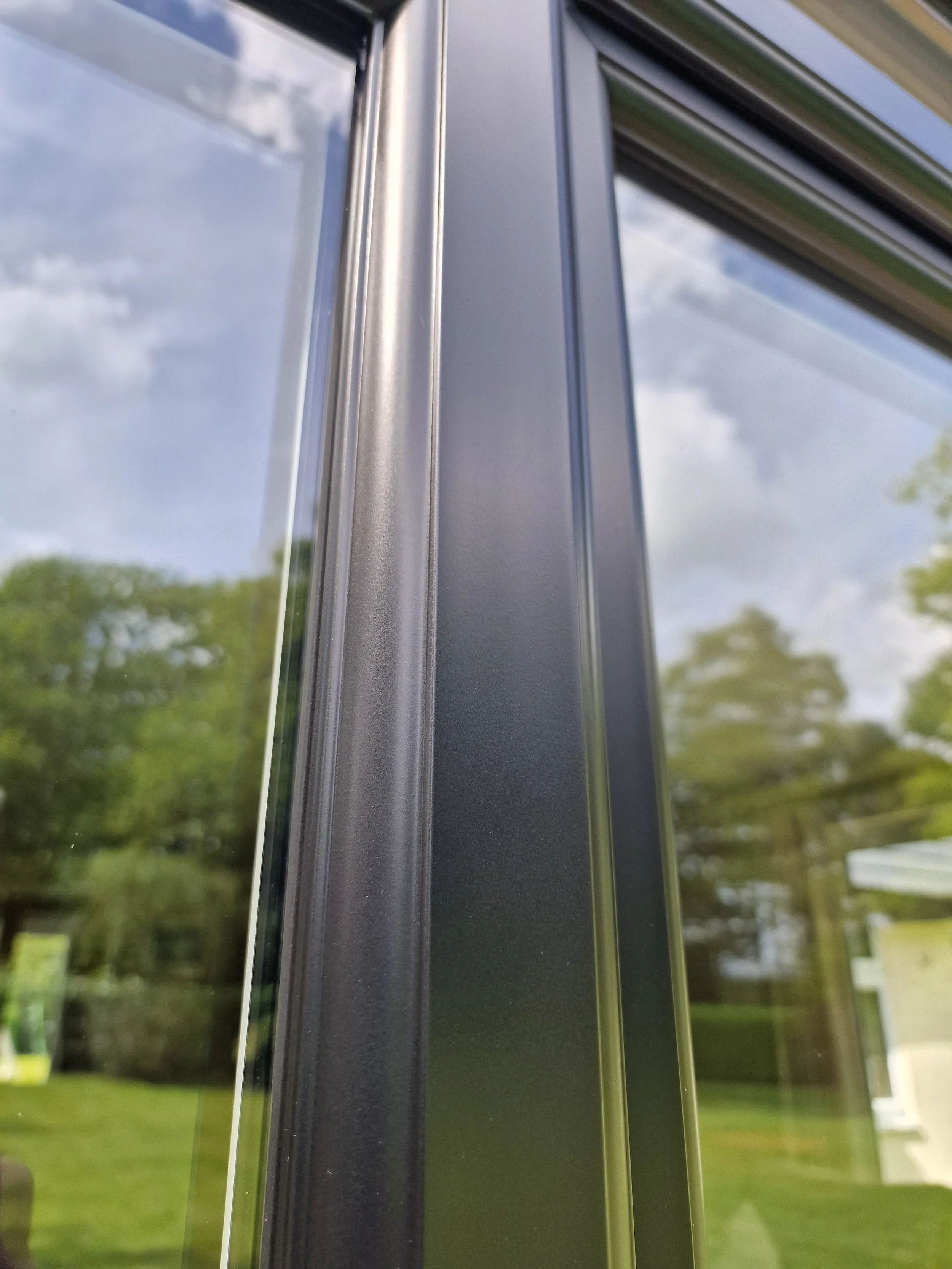 Close-up image of the metal frame of a window or door with reflective glass, showing trees and a cloudy sky outside.