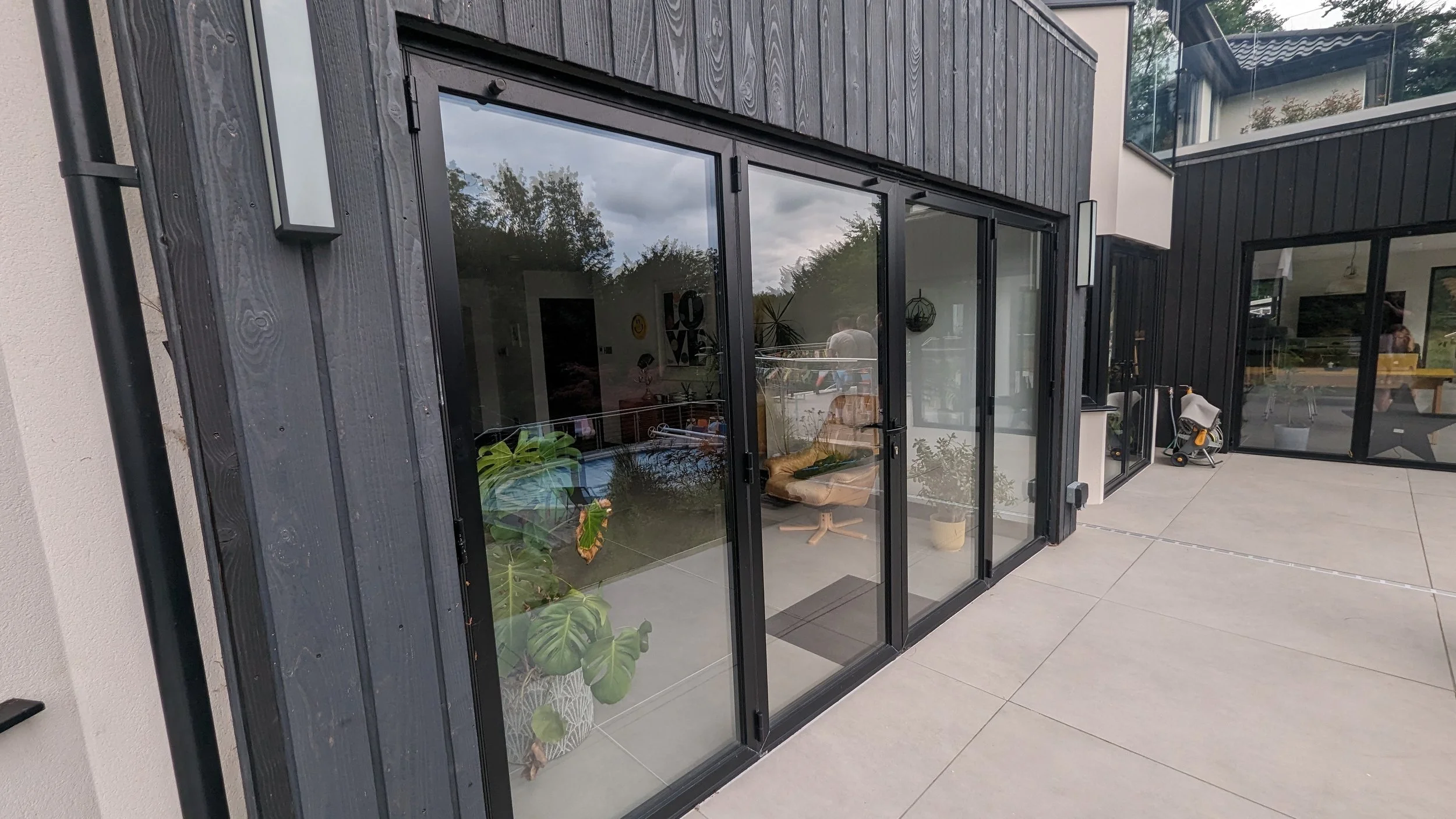 Modern house with large glass sliding doors, black wooden siding, outdoor patio with potted plants, and some children's toys.