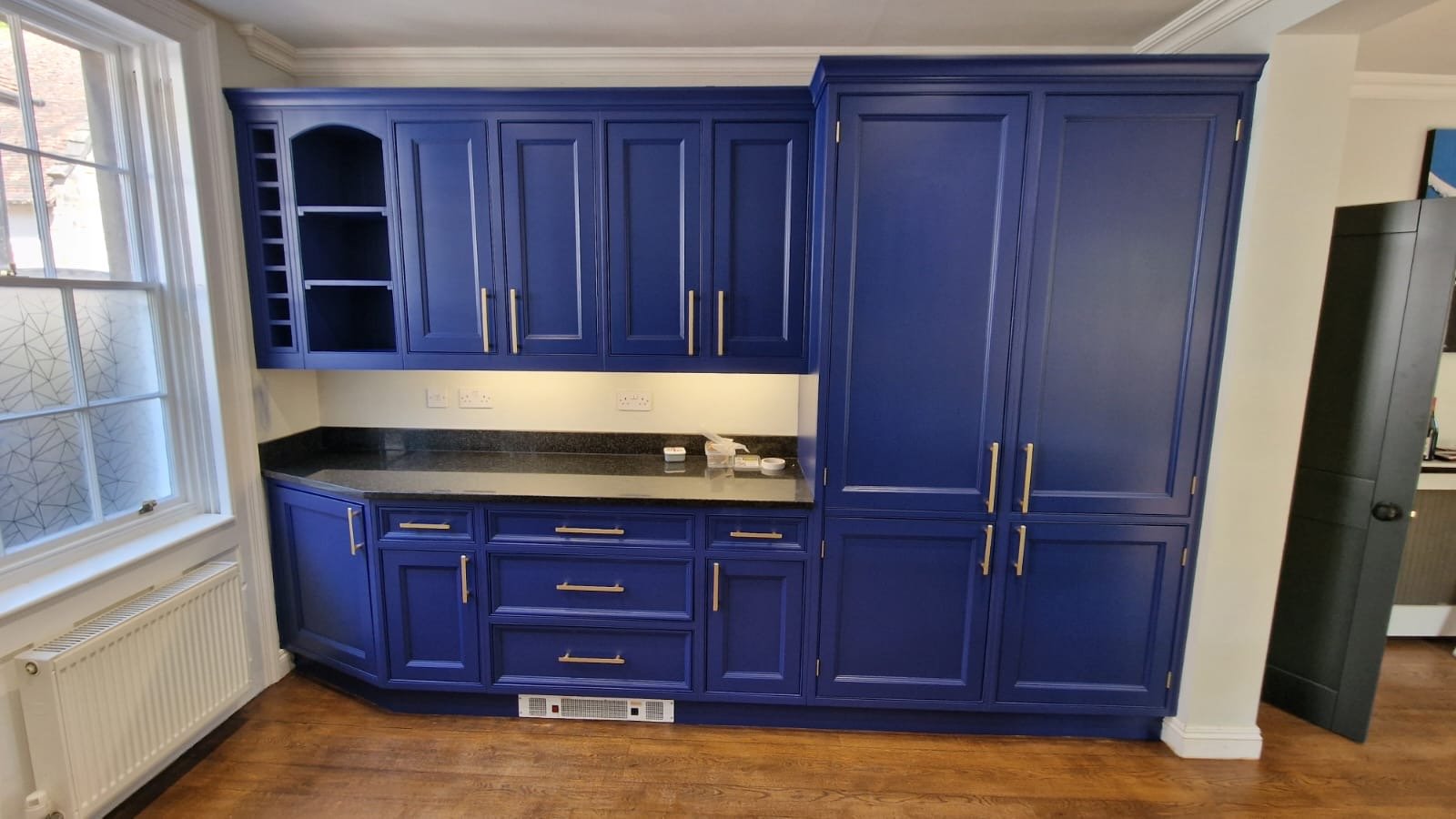 Blue kitchen cabinets with gold handles and black countertop, located near a window with white trim and wood flooring.