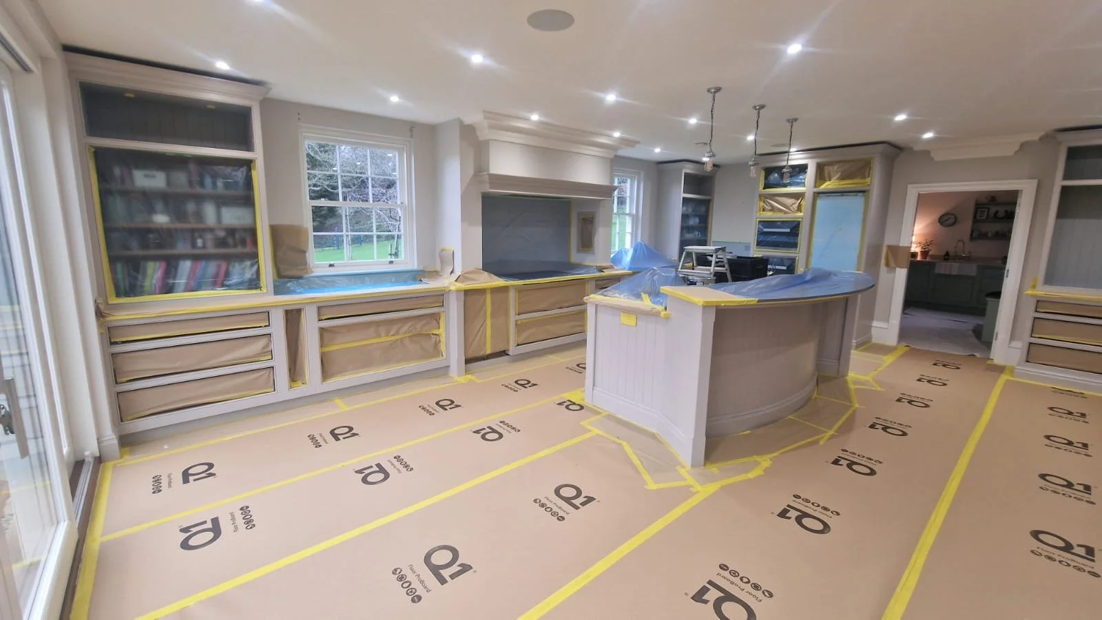 Kitchen under renovation with plastic-covered cabinetry, large windows, and safety tape on the floor indicating unfinished flooring.