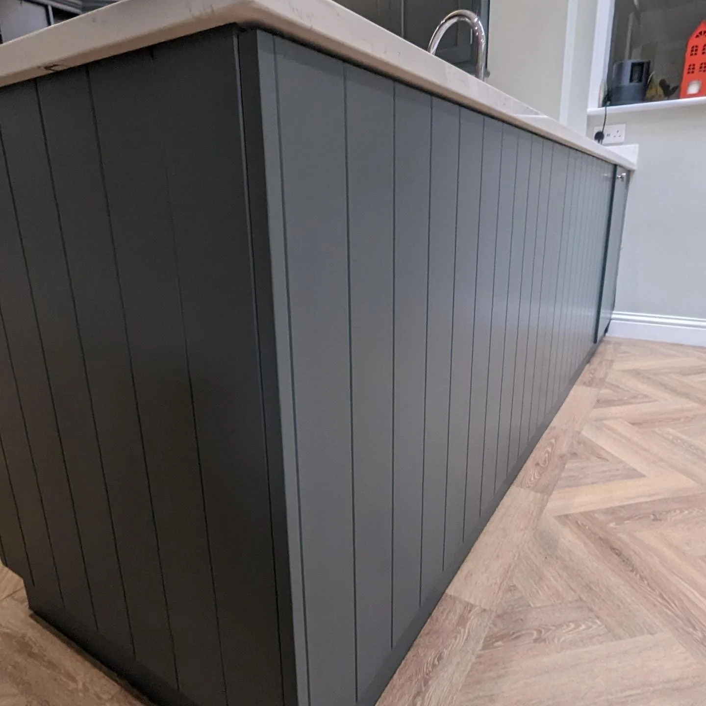 A kitchen island with grey wooden vertical paneling and a beige countertop. The island has a faucet and sink, located in a modern kitchen setting with hardwood flooring and a shelf with various items in the background.