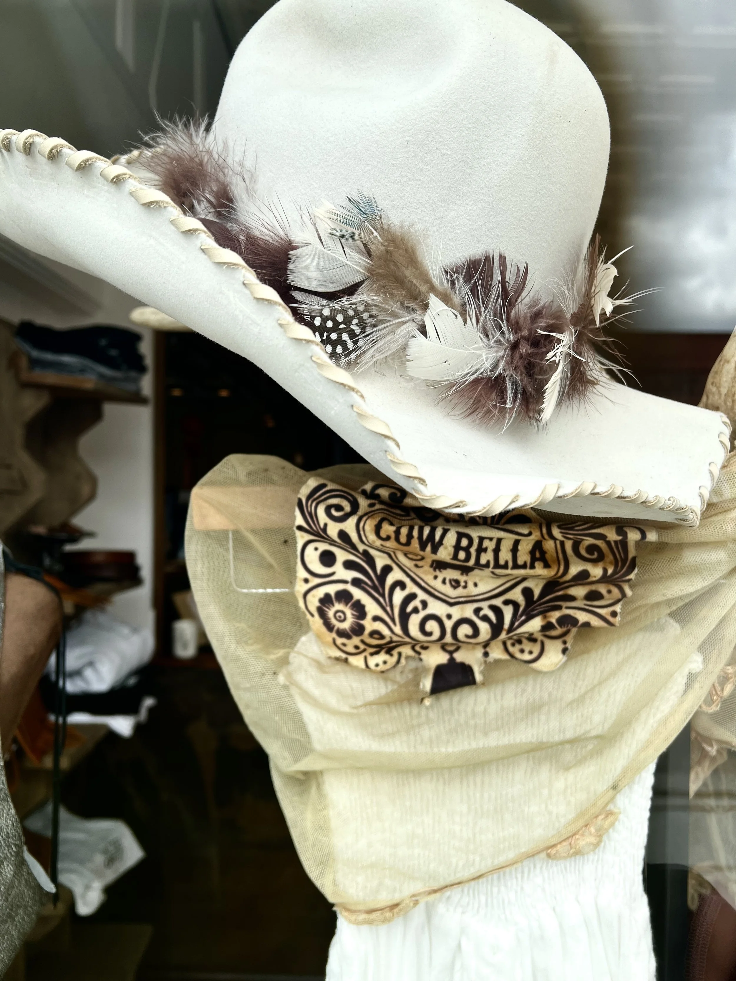 A white cowboy hat decorated with multiple feathers, resting on a beige cowboy hat with a decorative band that reads 'COW BELLA'.