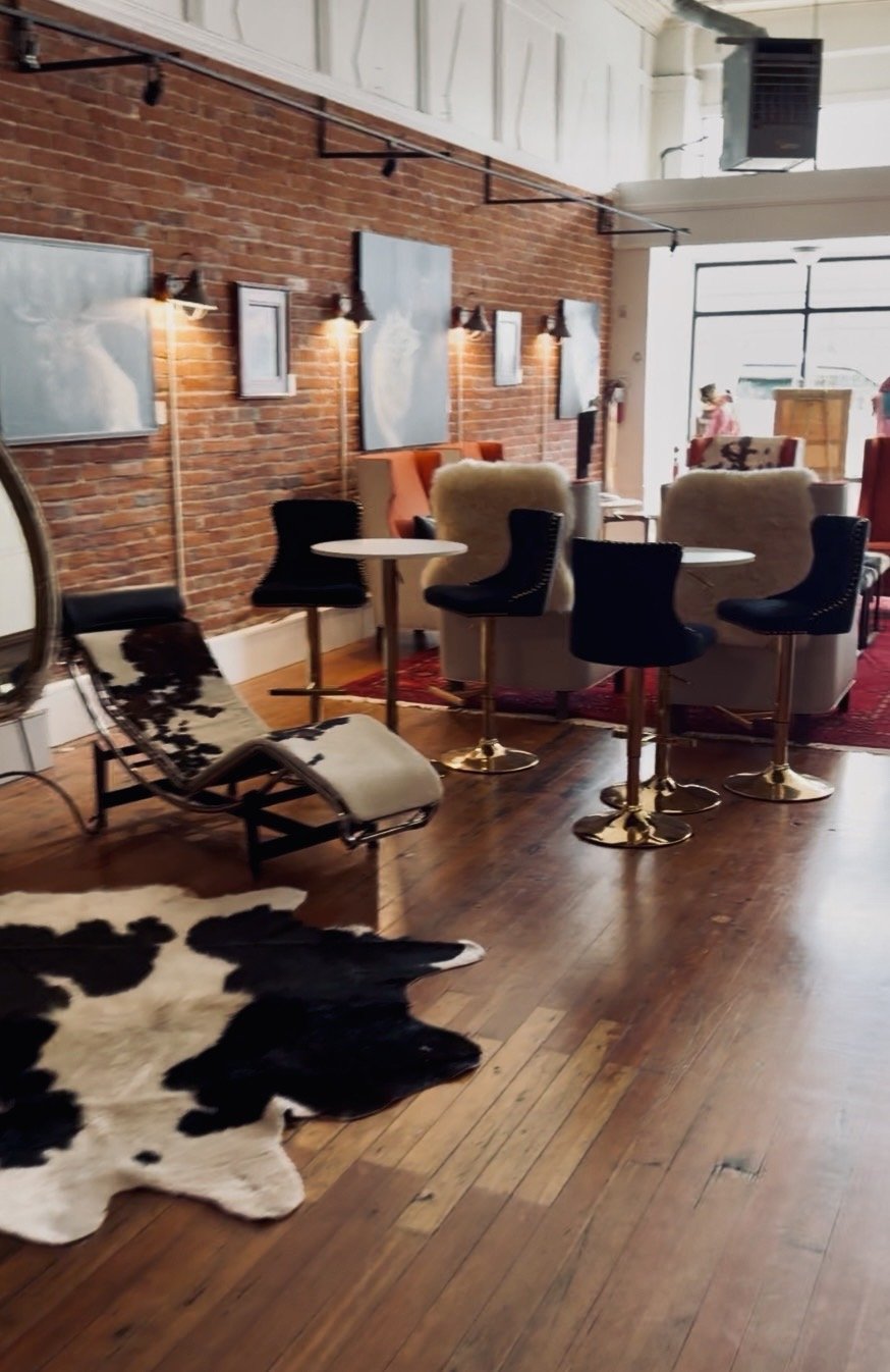Interior of a cozy cafe or lounge with brick wall, wooden floors, modern black chairs around small tables, a cowhide rug on the floor, and artwork on the wall. Sunlight filters through large windows at the back.
