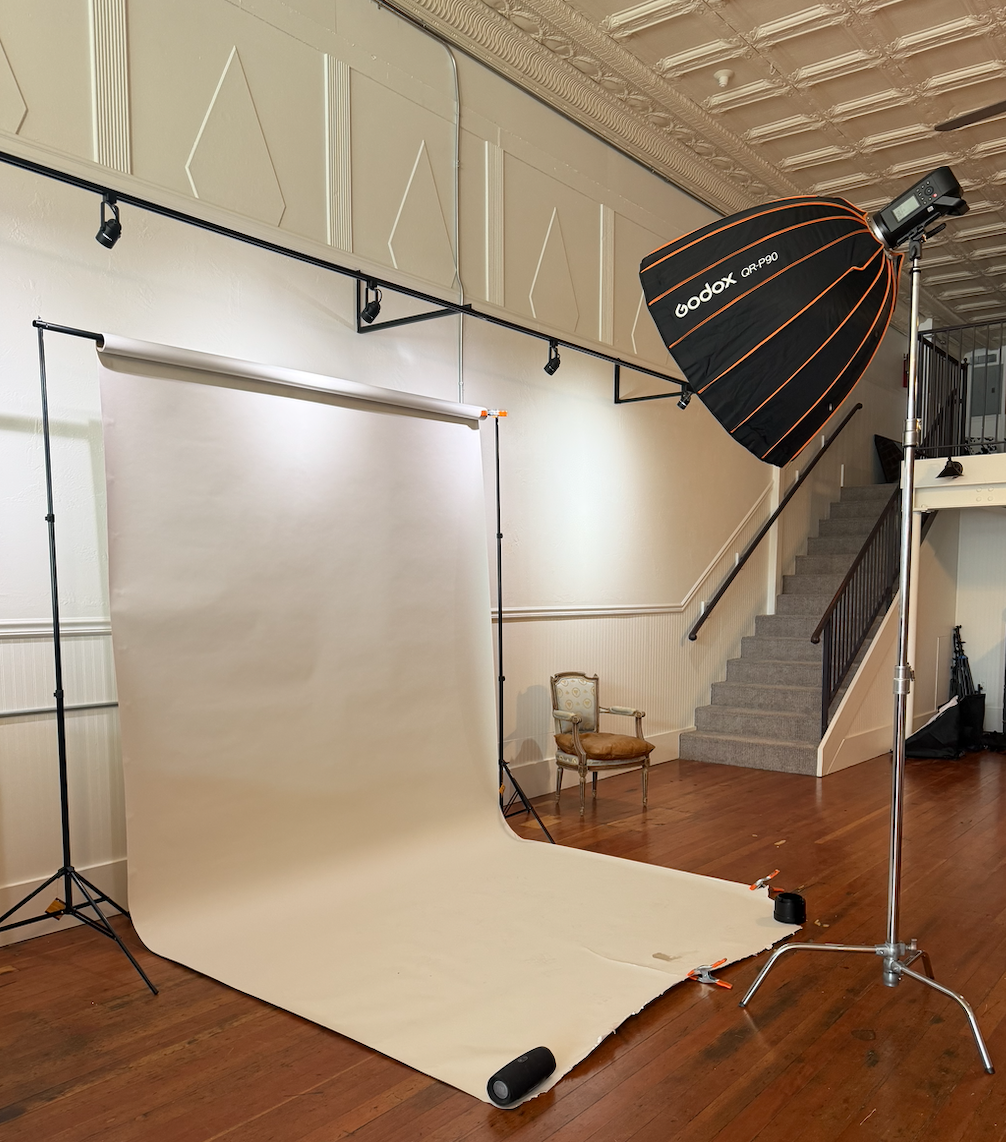 Photography studio setup with a white backdrop, a large softbox light, and a chair on a staircase landing. Wood floor and decorative ceiling.