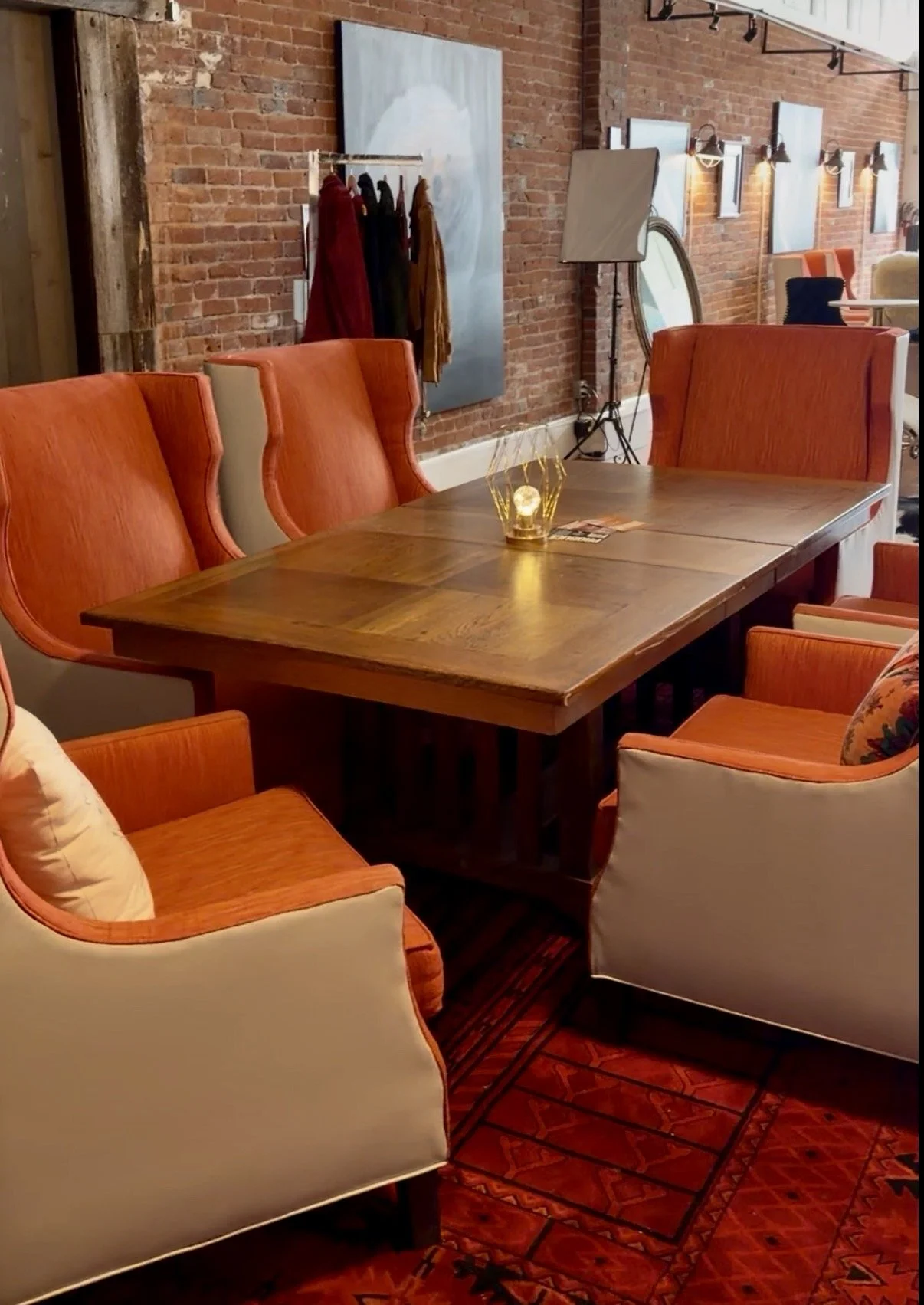 A cozy dining area with a large wood table surrounded by orange and white upholstered chairs, brick walls, and a decorative rug.