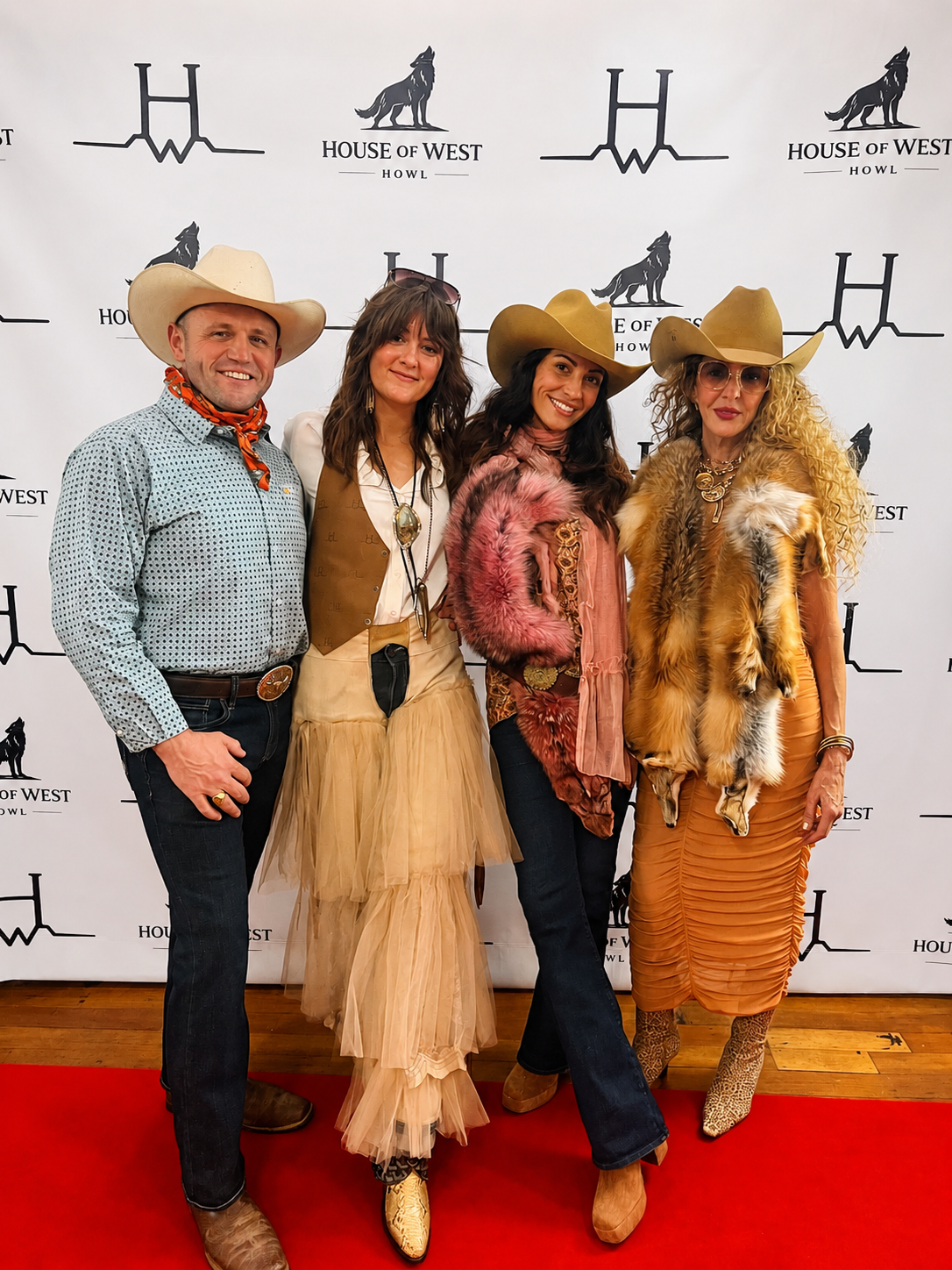 Four people dressed in western cowboy attire, standing on a red carpet in front of a white backdrop with 'House of West' and a wolf logo, smiling for the photo.