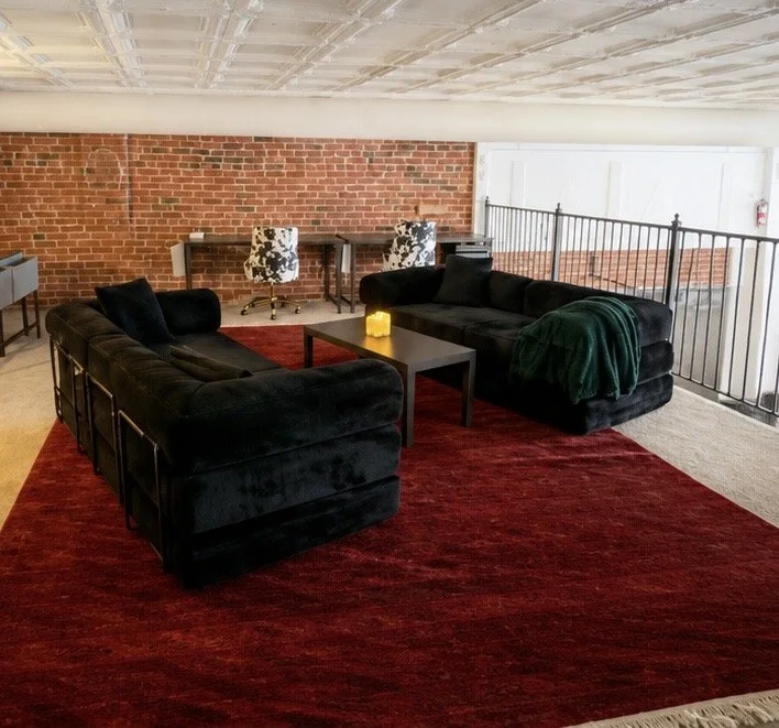 Living room area with two black sofas on a red rug, a black coffee table with a yellow candle, a brick wall, and a small private balcony with railings. In the background, there is a desk with two patterned chairs.
