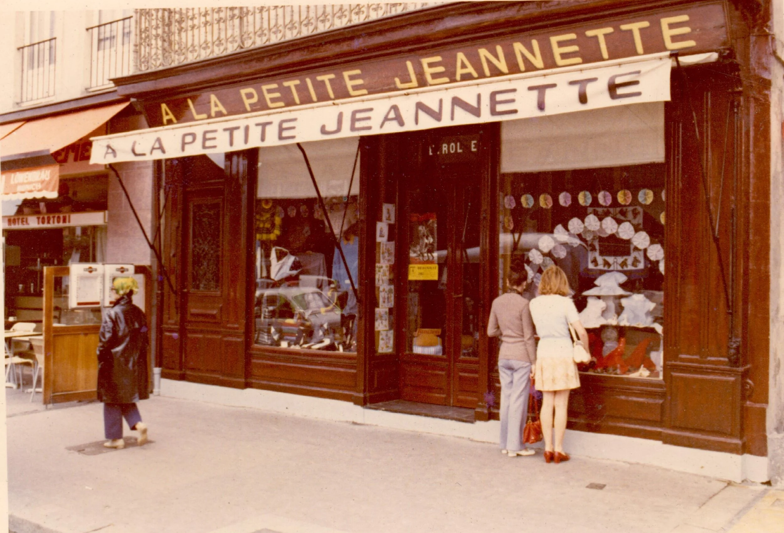 Façade de la boutique historique en bois avec l'enseigne annonçant 'À La Petite Jeannette'. Trois personnes, deux femmes et un homme, regardent la vitrine, avec des cachemires et des décorations colorées visibles à l'intérieur.