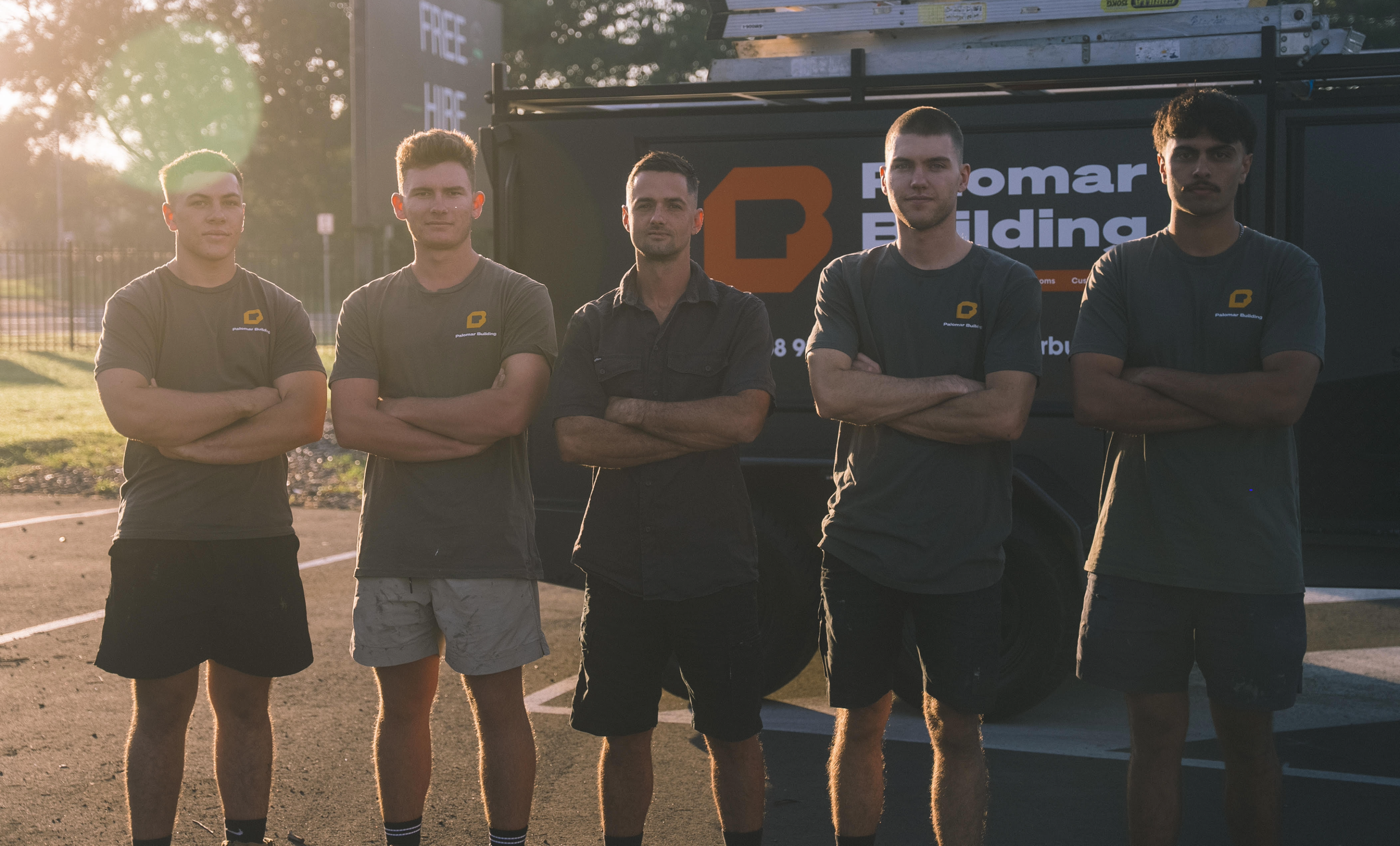 Five young men standing with arms crossed in front of a Palomar Building truck, outdoors during sunset.