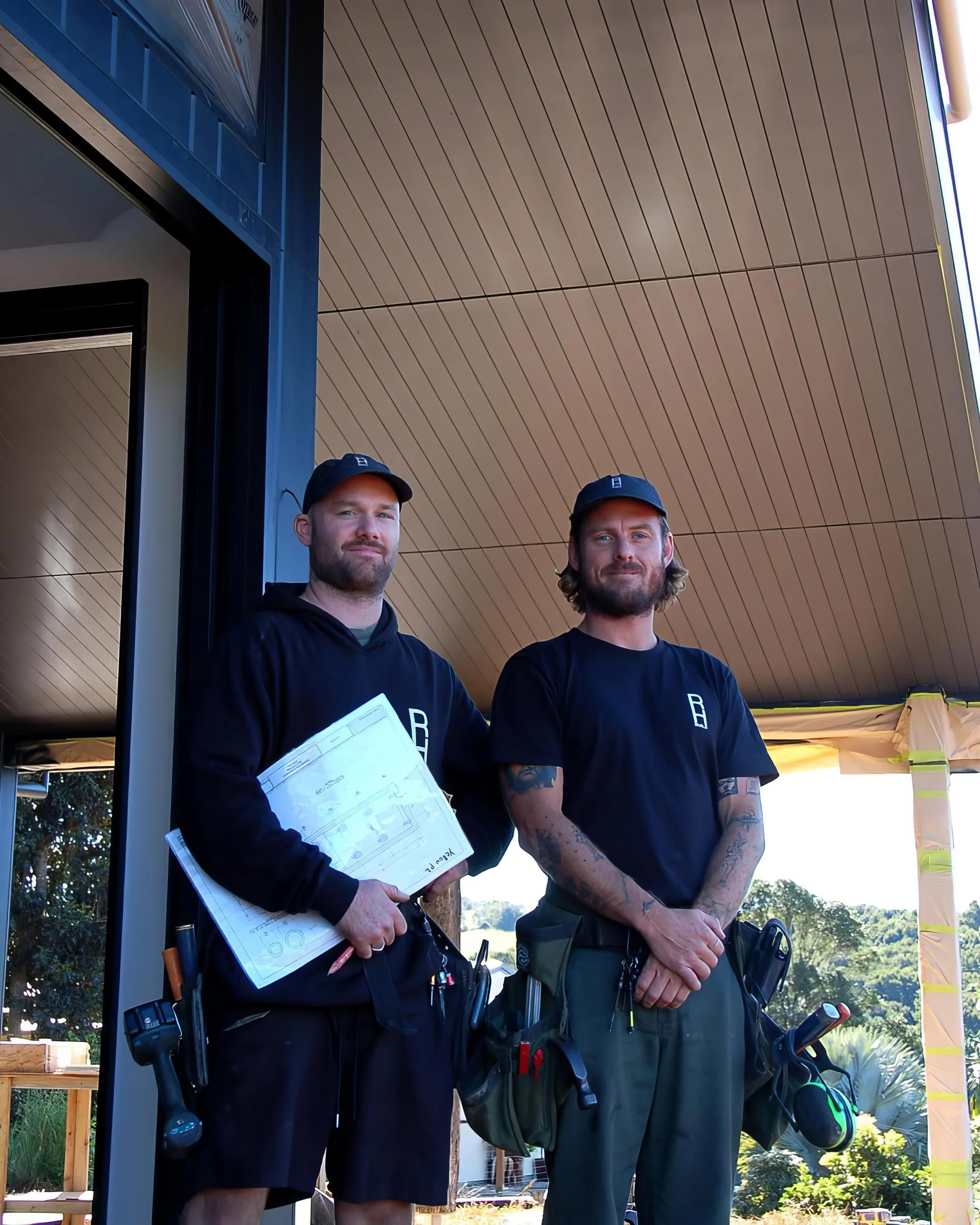 Two male construction workers standing outdoors under a wooden overhang, wearing black shirts, black caps, and tool belts, with a sunny background of trees.