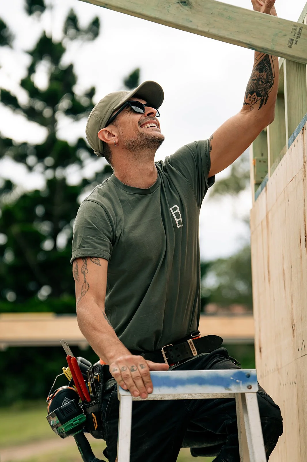 Man smiling and working on a construction project outdoors, wearing a hat, sunglasses, and a tool belt, holding a ladder for support.