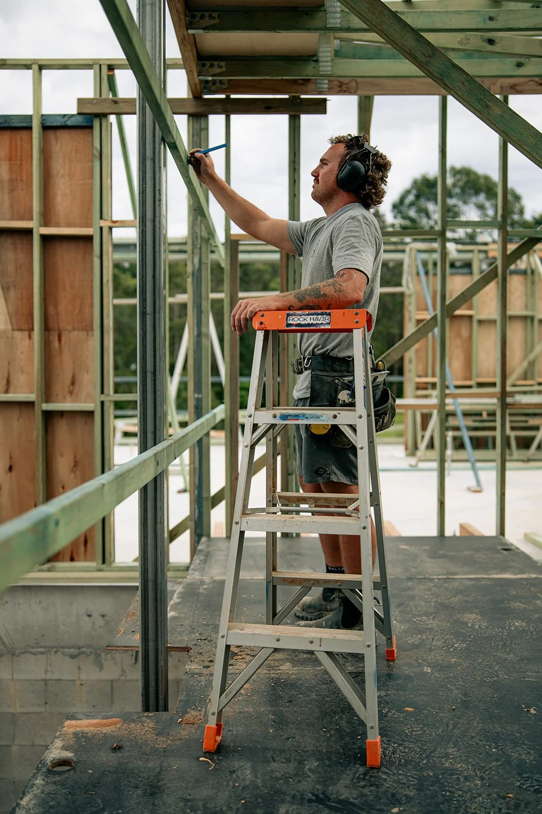 A man on a ladder working on the framing of a building under construction, wearing hearing protection, a grey t-shirt, and shorts.