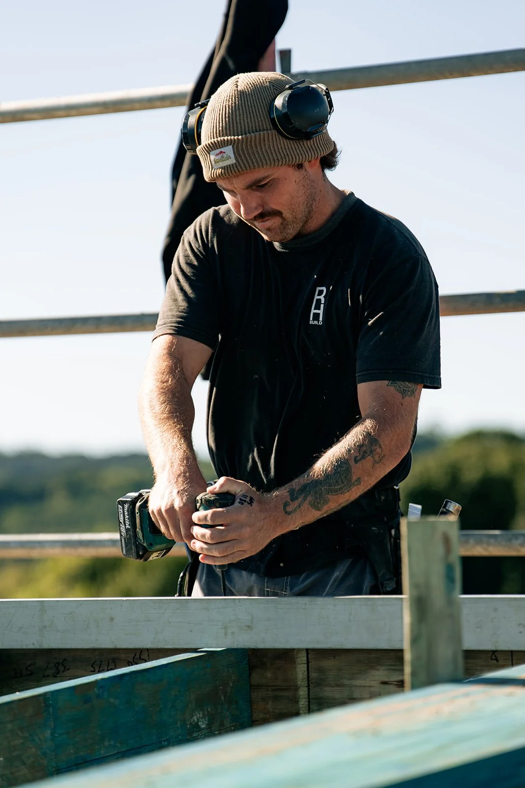 A man wearing a tan beanie, black t-shirt, and headphones around his neck is working with a cordless drill on a wooden structure outdoors.