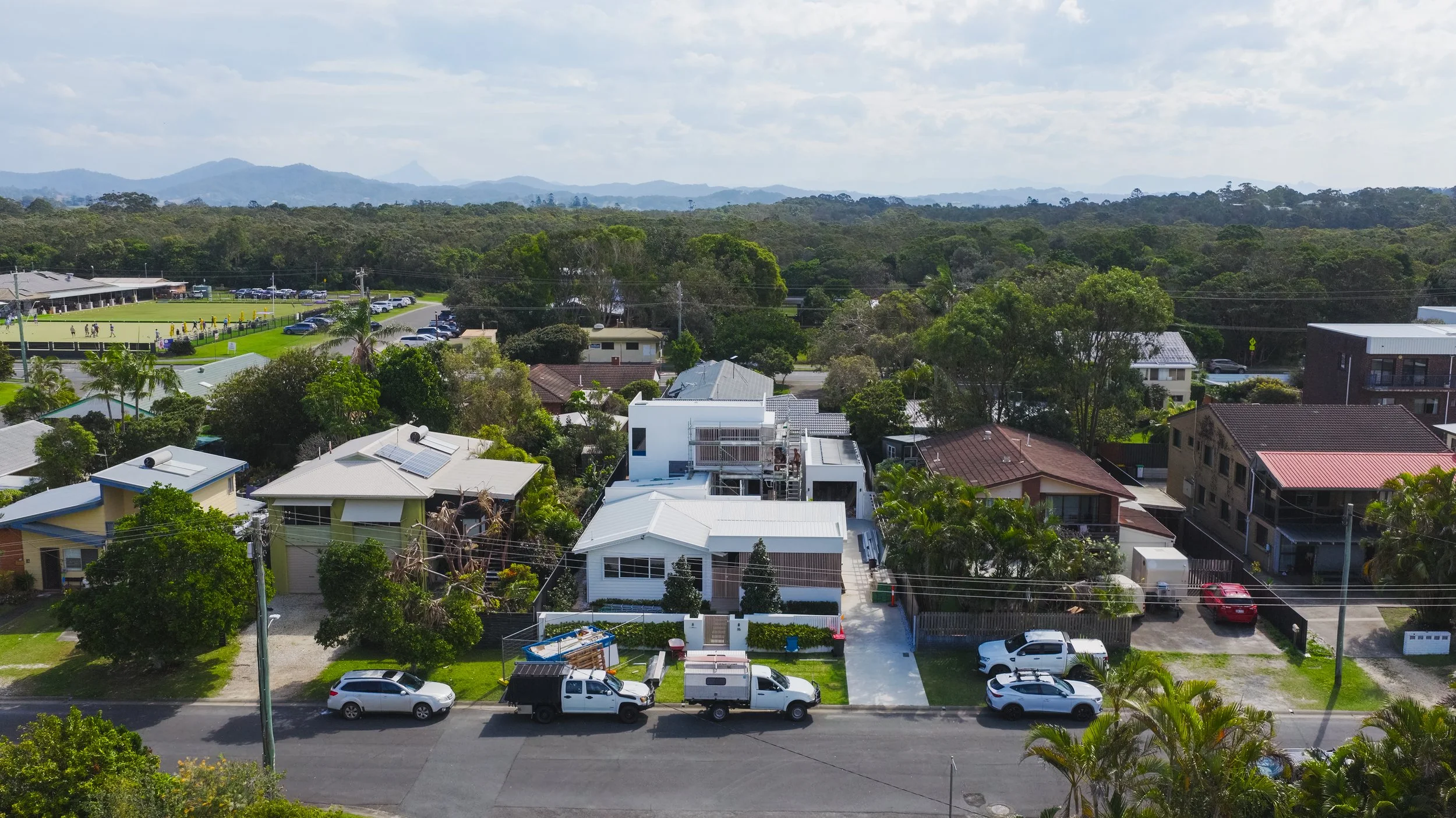 An aerial view of a suburban neighborhood with modern houses, parked vehicles, lush trees, and a sports field in the background, with mountains in the distance under a partly cloudy sky.