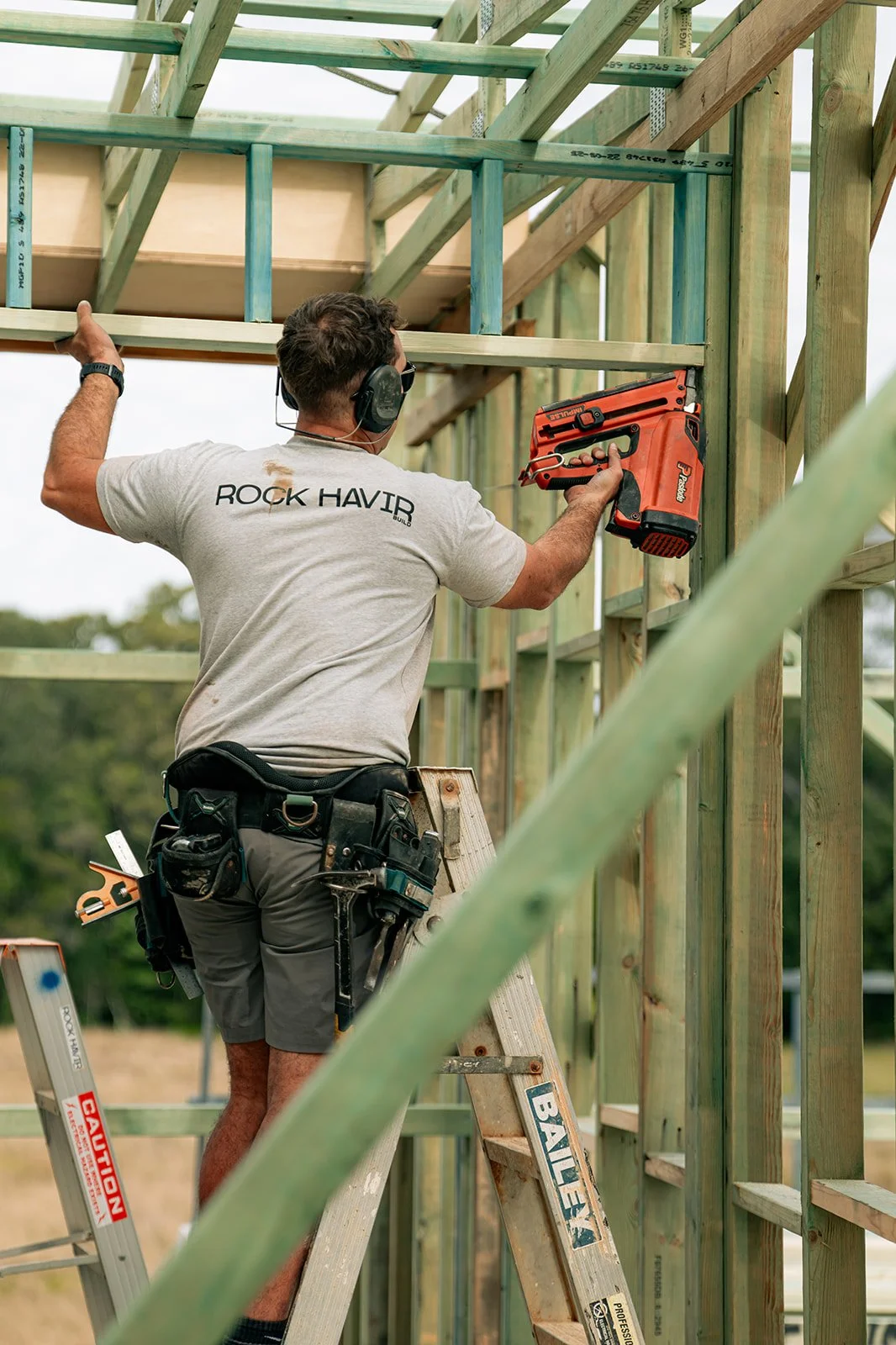 A man working on a wooden building frame uses a nail gun, wearing a tool belt, shorts, a gray T-shirt, and work headphones, standing on a step ladder outdoors.