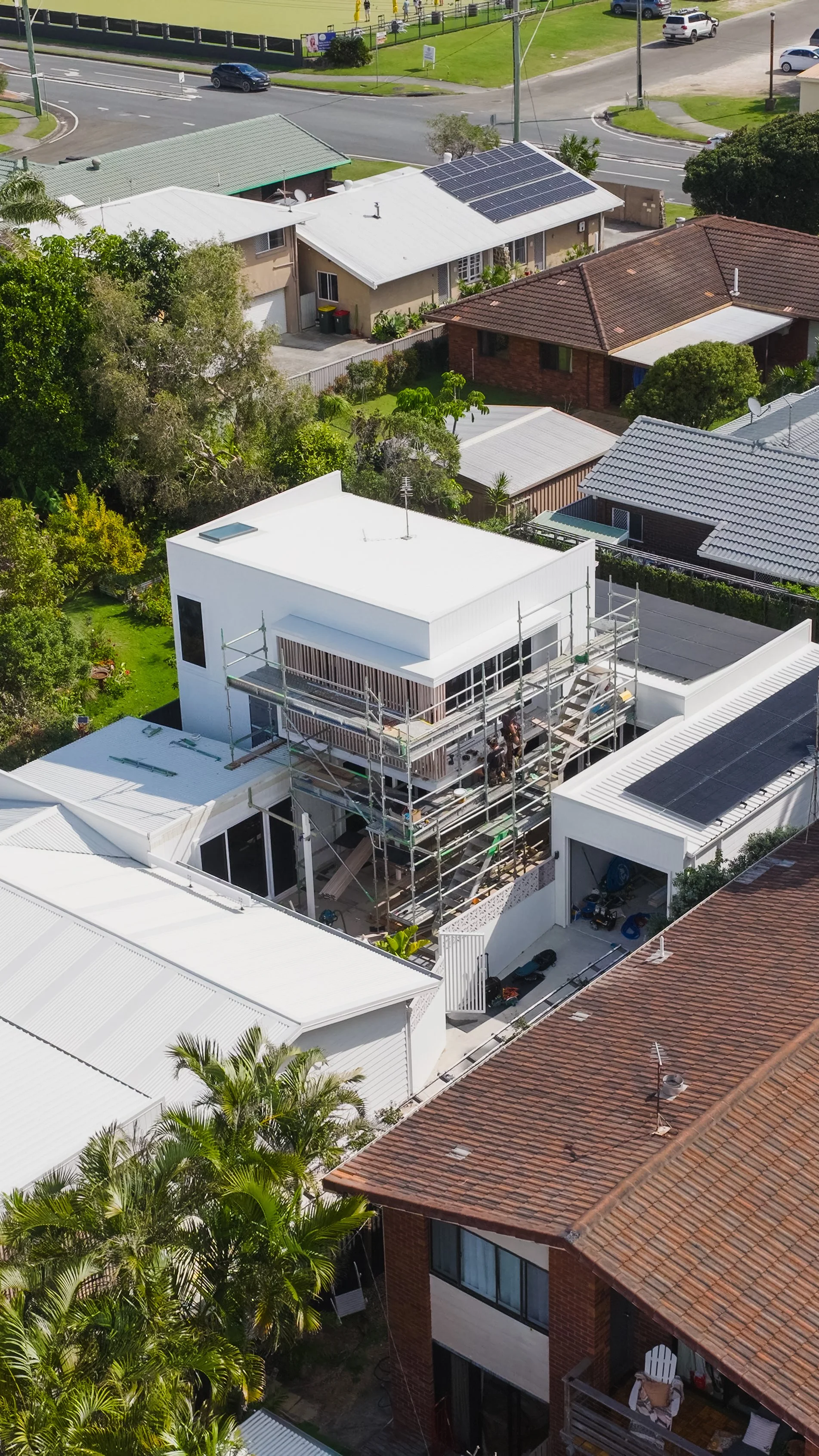 Aerial view of a residential neighborhood showing houses with various roof styles, including one under construction with scaffolding, solar panels on some roofs, and lush green trees and gardens.