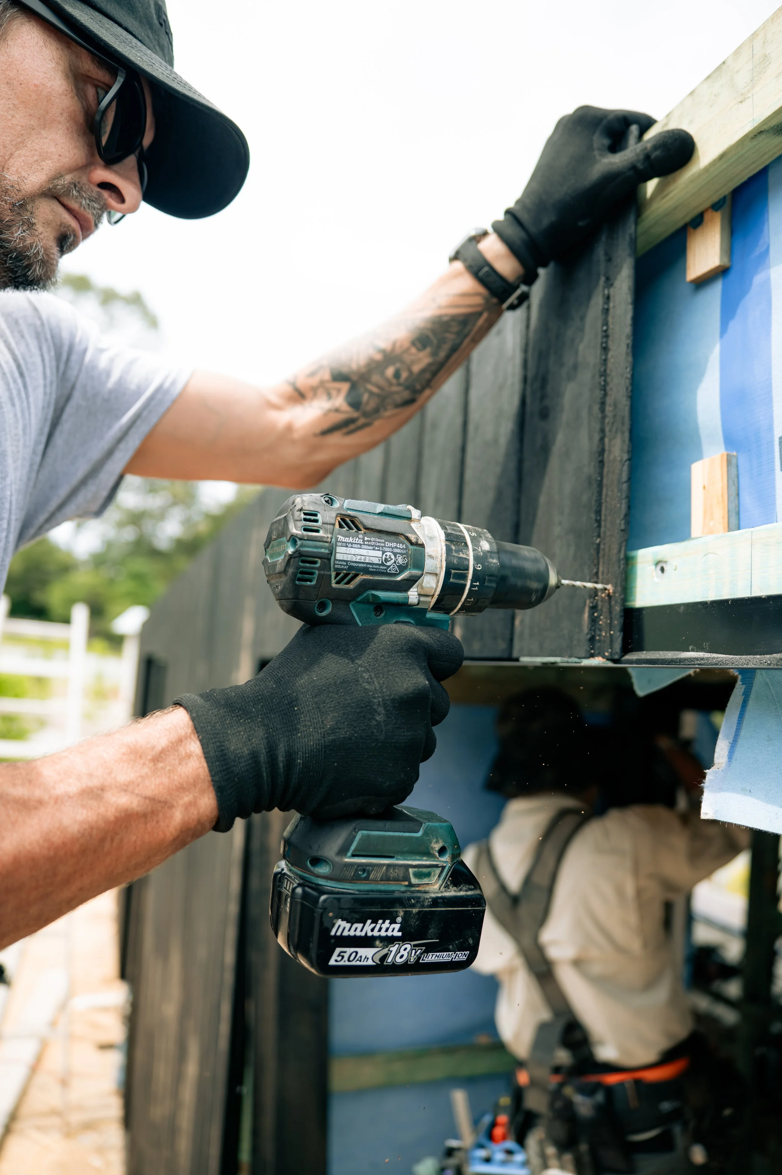 A man using a cordless drill to secure a piece of wood to an outdoor structure, with another person working underneath.