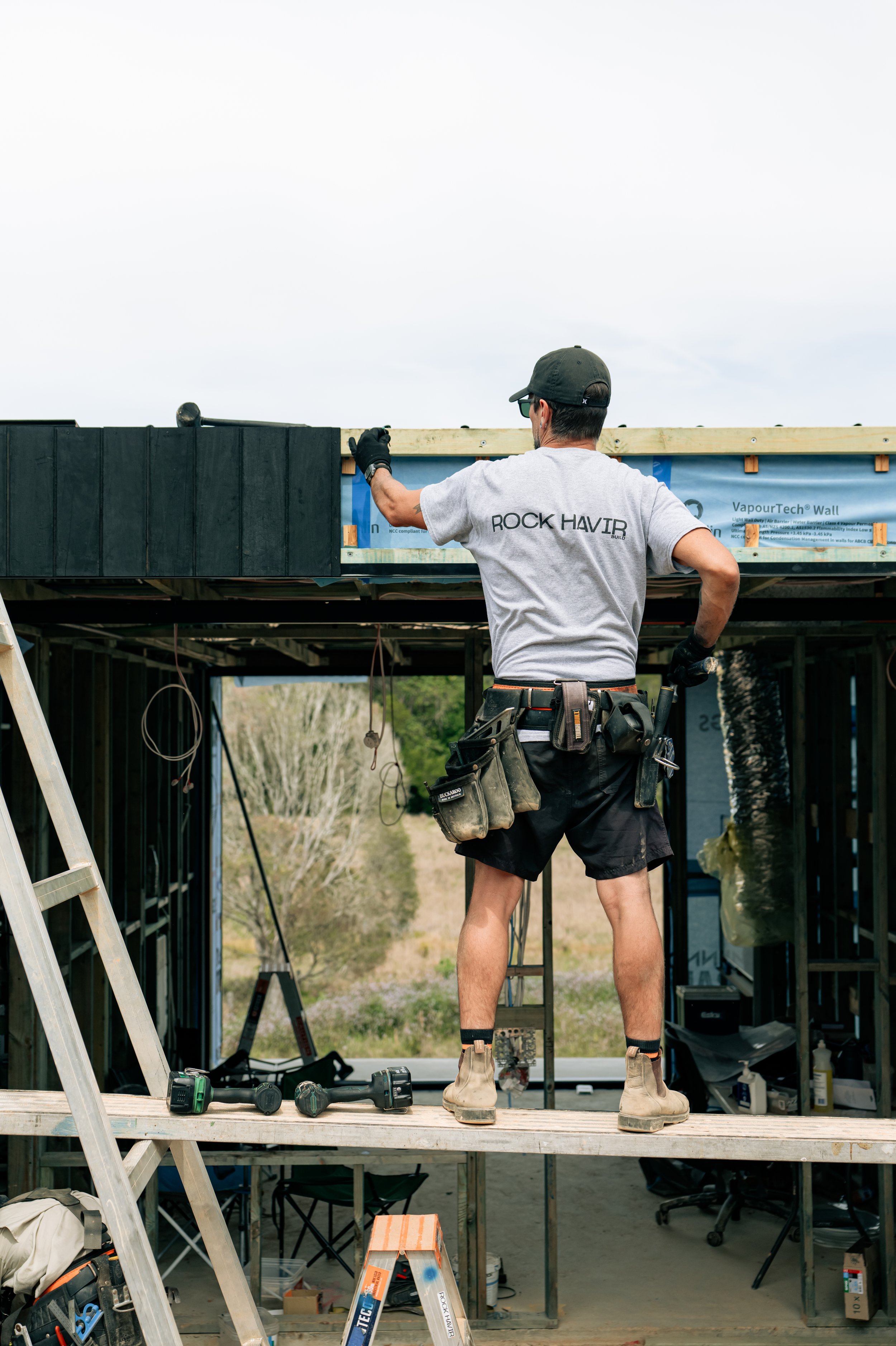 A man working on a construction site, standing on a wooden platform, installing a roof or wall. He is wearing a gray t-shirt, black shorts, gloves, and a tool belt, with a ladder and construction tools nearby.