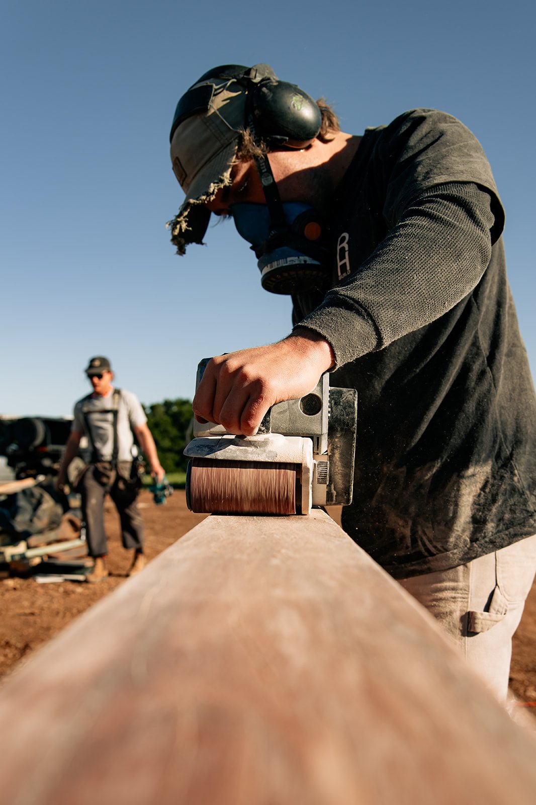 A person sanding a wooden beam with a power sander, wearing protective gear including a helmet, ear protection, and a mask, with another person in the background.