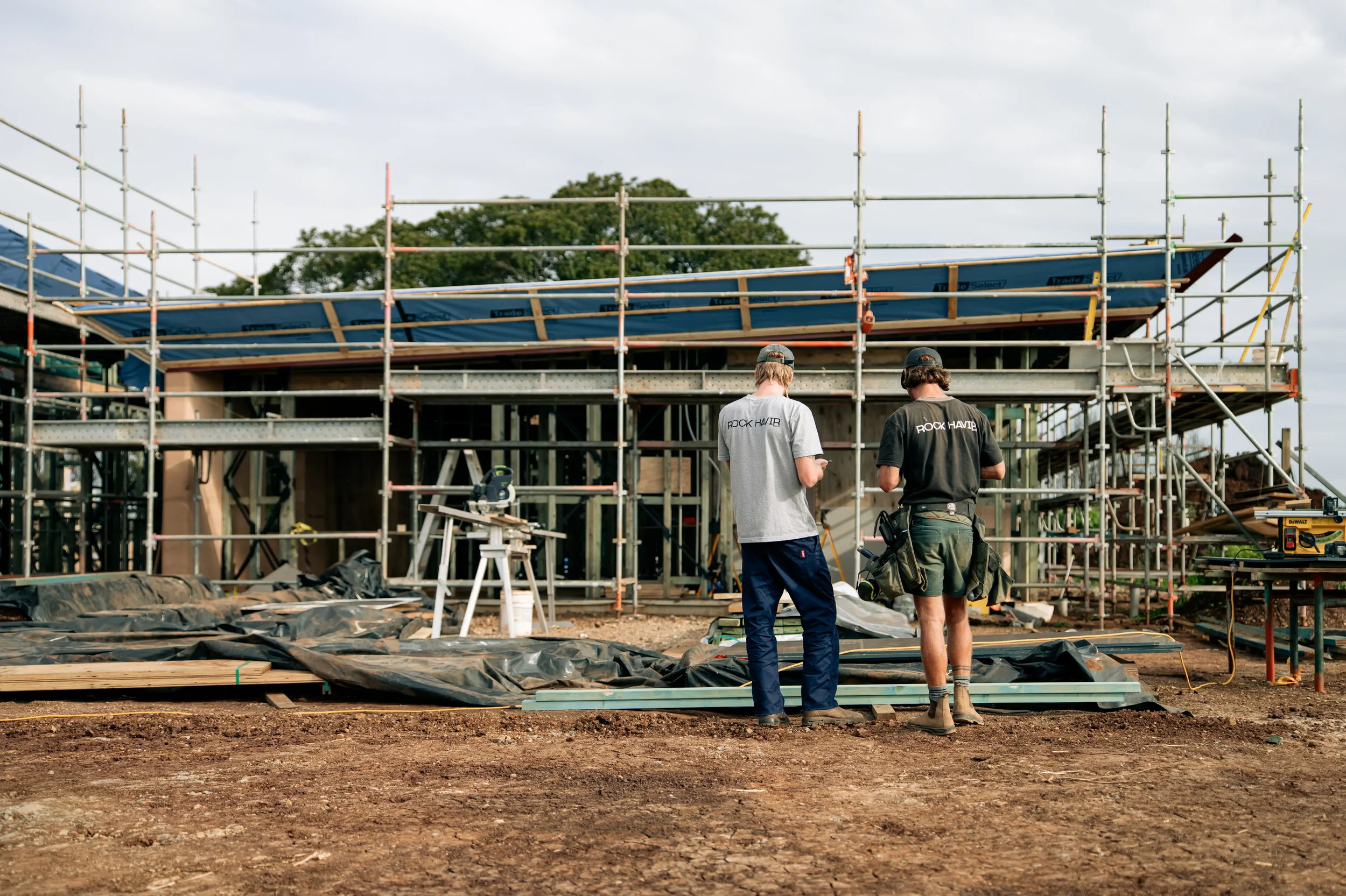 Two construction workers stand in front of a building under construction, surrounded by scaffolding, with construction tools and materials on the ground.