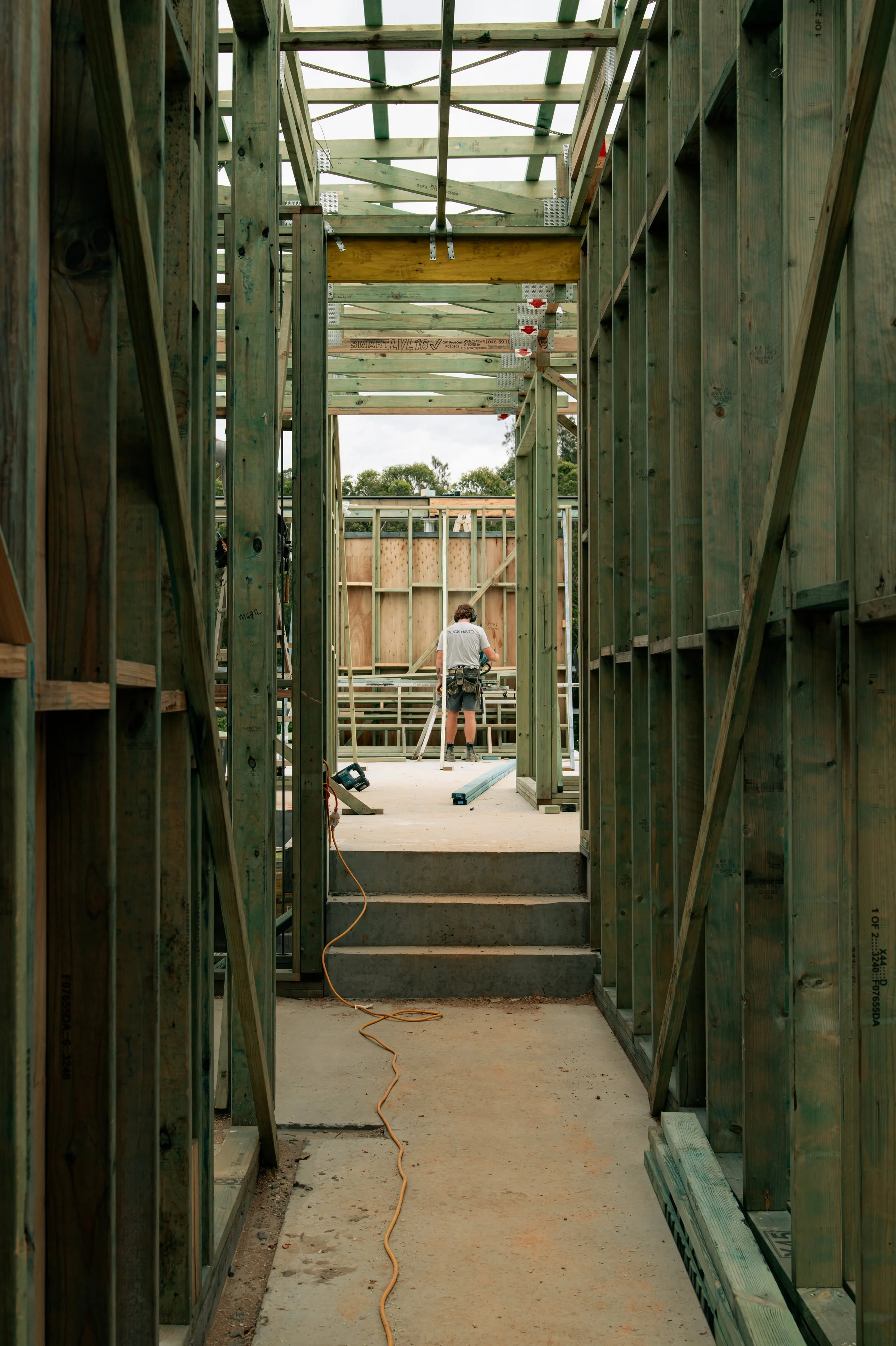 Construction worker building a house, standing on a wooden platform, surrounded by wooden framing and stairs, with tools and construction materials around.