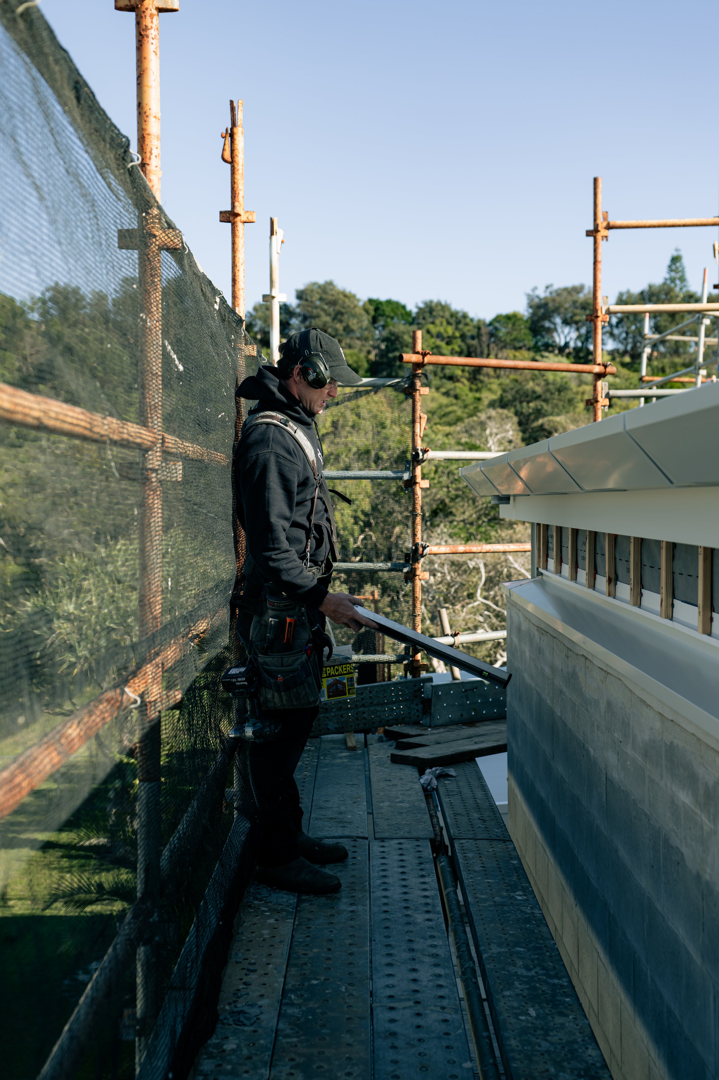 A construction worker standing on scaffolding, wearing a black hoodie, black cap, ear protection, and holding a tool, working on the exterior of a building with a white roof. The background shows a hillside with green trees under a blue sky.