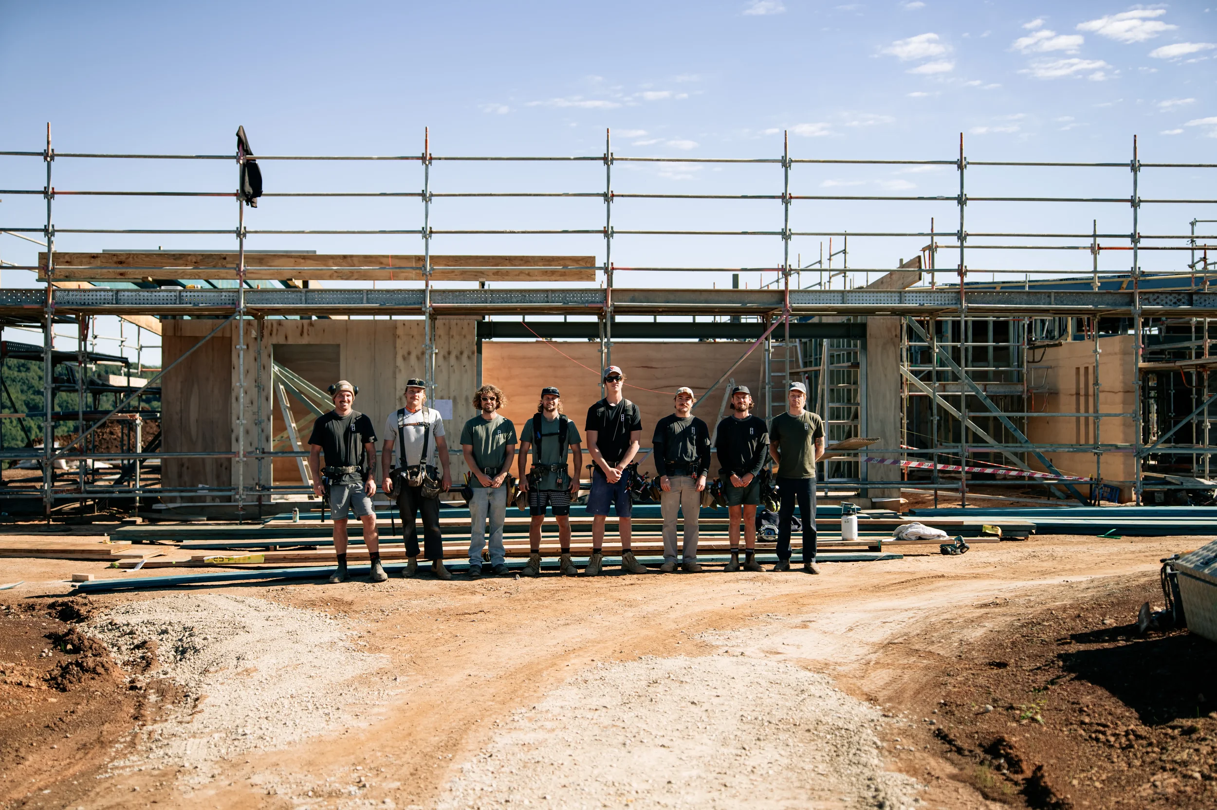Group of nine construction workers standing in front of a partially built wooden structure at a construction site with scaffolding and blue sky.