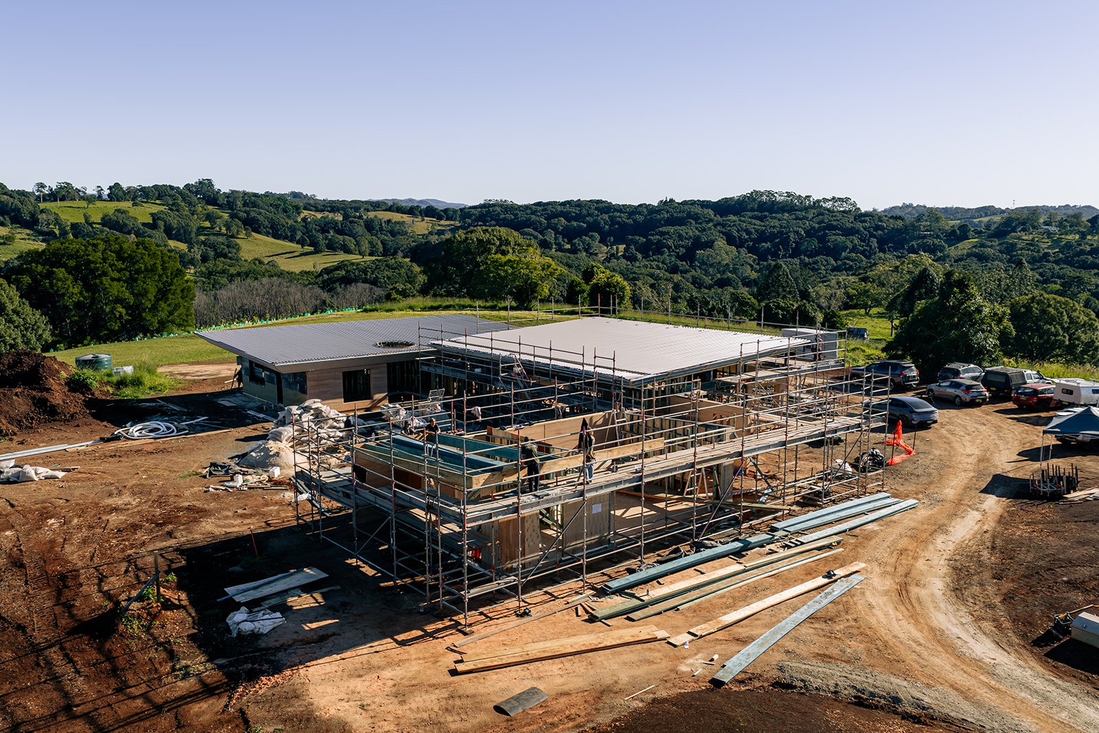 Construction site with a partially built house surrounded by scaffolding, with a scenic landscape of green rolling hills in the background under a clear blue sky.