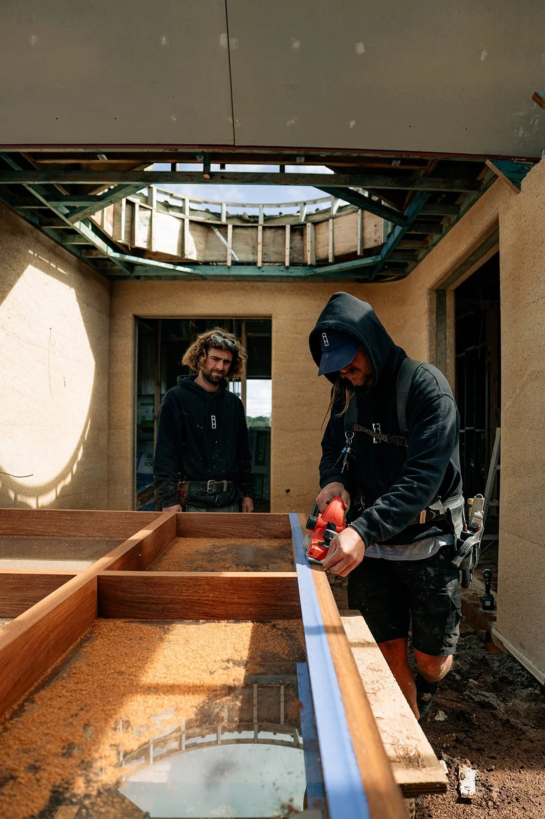 Two construction workers, one cutting wood with a power saw, at a house under construction, with a partially built roof and an open sky above.